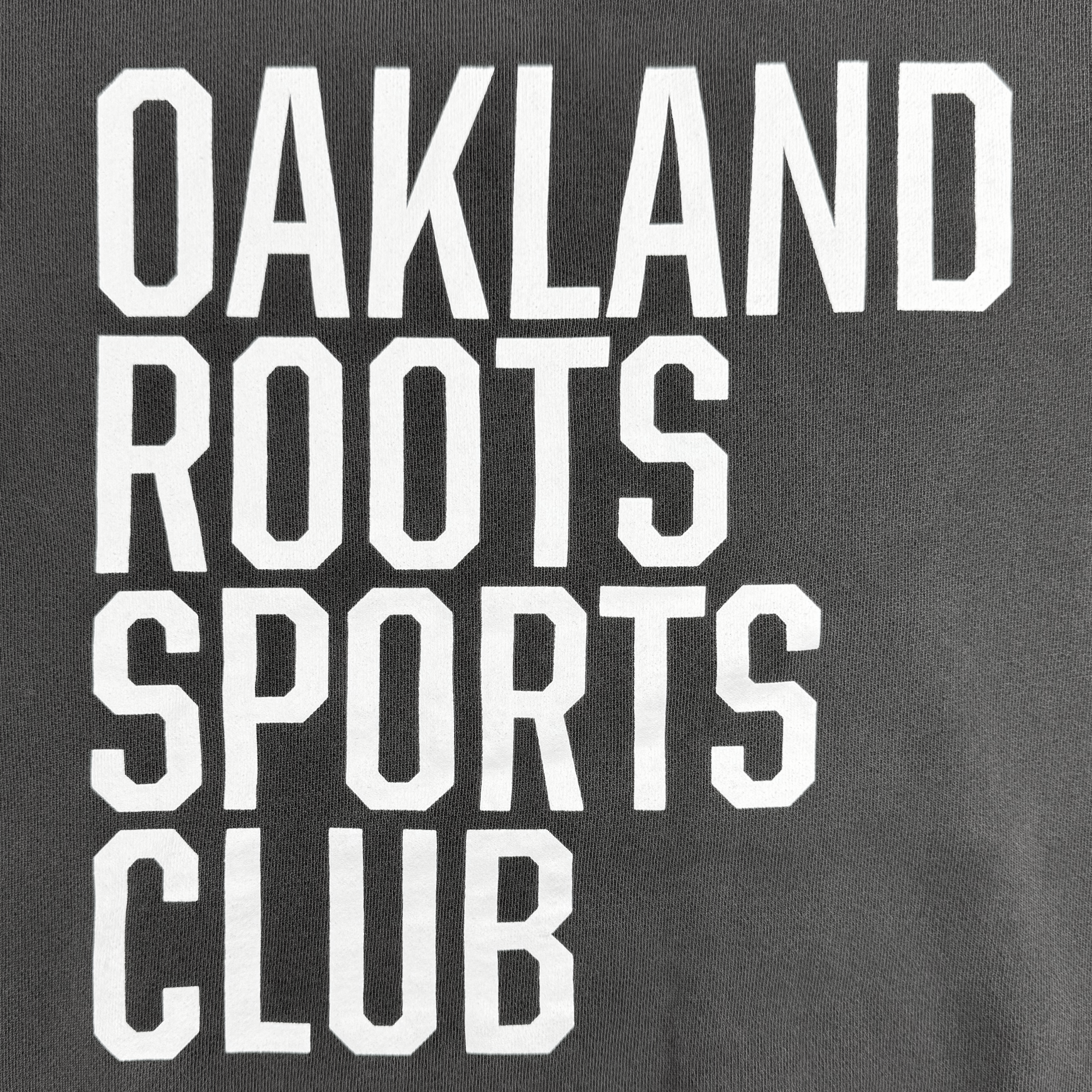 The image showcases a dark gray backdrop adorned with bold, uppercase white lettering organized in four lines, prominently displaying the "OAKLAND ROOTS SC" name. The heavy and modern font underscores Oakland Roots SC's dedication to social good and community engagement.