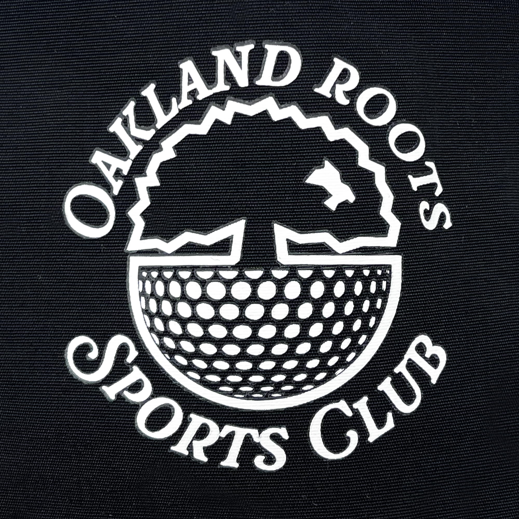 The Heritage Bucket Hat by Oakland Roots SC features a black background adorned with a white emblem of the Oakland Roots Sports Club. The emblem showcases a stylized tree with roots in the pattern of a soccer ball at the bottom. "Oakland Roots Sports Club" is written in a circular arrangement around this tree and roots design, representing their mission of social good.