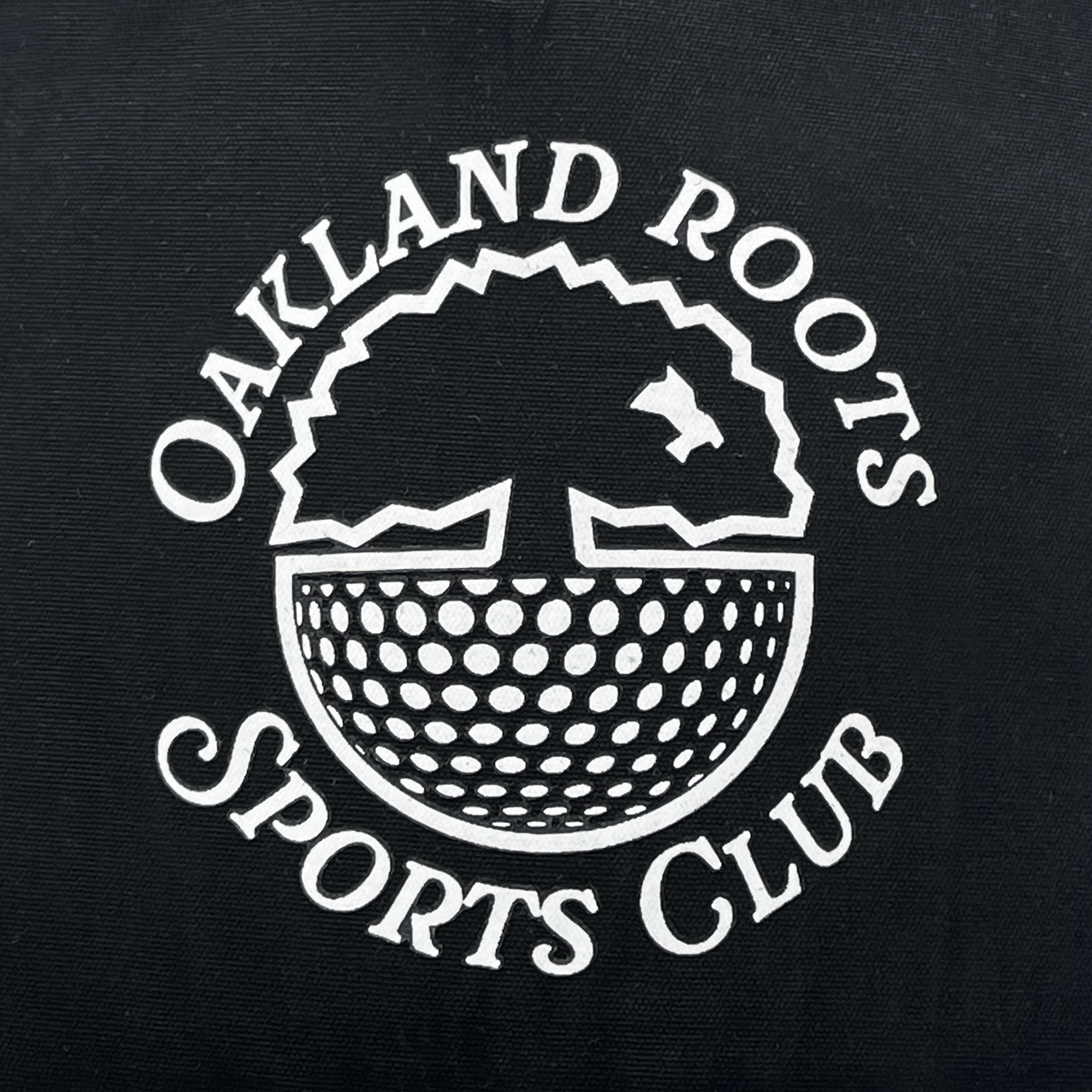 The image shows the logo of Oakland Roots SC on the Heritage Cap. The logo features "Oakland Roots SC" encircling a stylized semi-circular tree, with its crown forming an upward arch and its roots as lines extending downward.