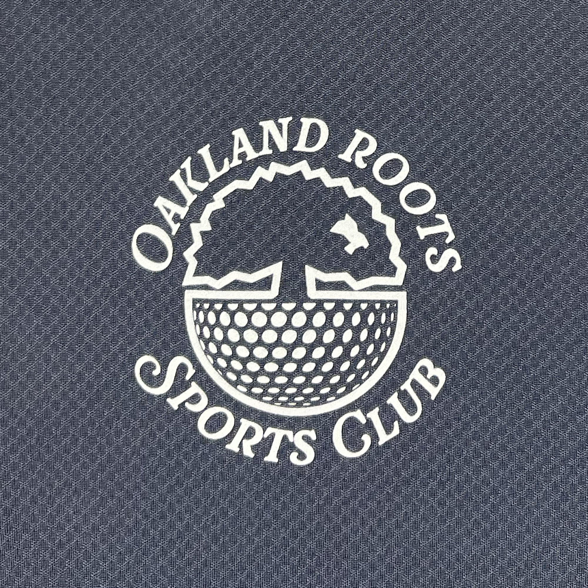 Logo of the Heritage Polo by Oakland Roots SC with white text and graphic on a textured gray background. The logo features a tree with roots forming a soccer ball pattern, reminiscent of an Oaklandish Polo Tee design. A bird is flying behind the tree, and the words "Oakland Roots Sports Club" surround the graphic in a circular formation.