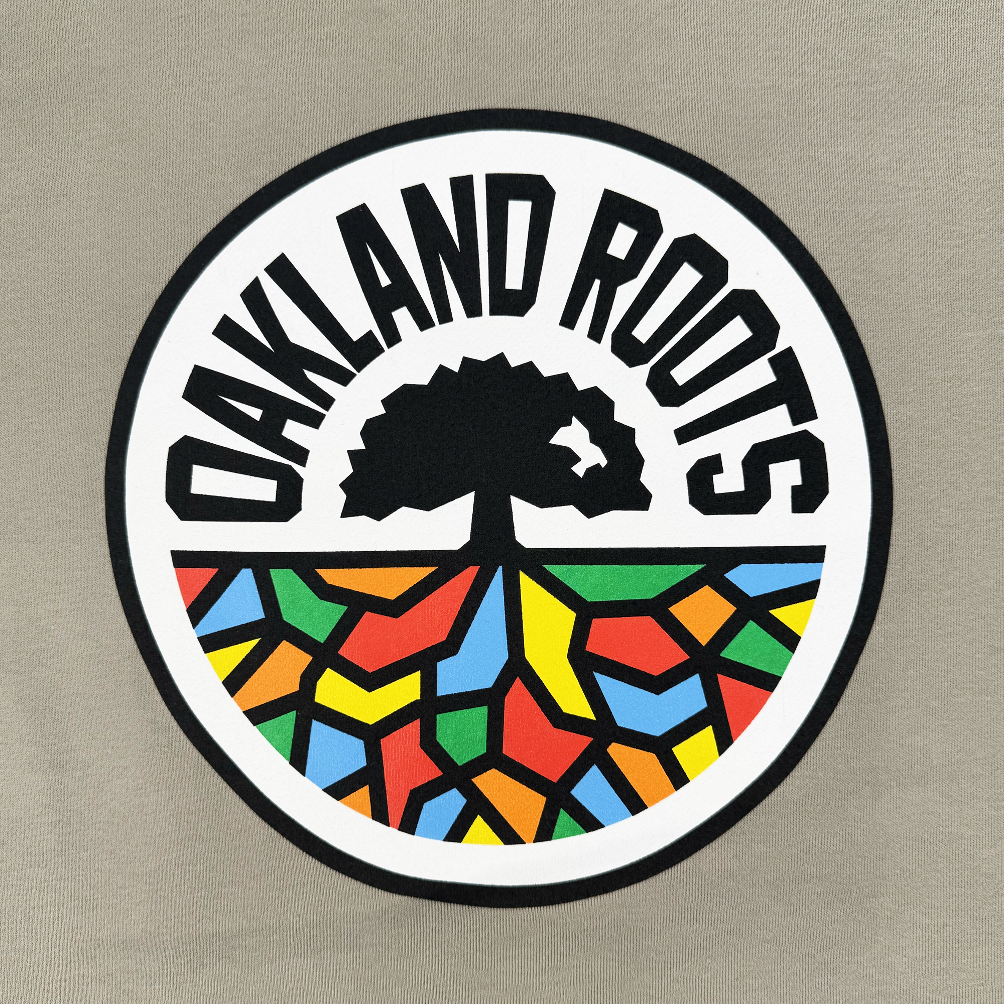 The Oakland Roots SC Classic Hoodie features a logo with "Oakland Roots SC & Soul" around a stylized tree. Below, vibrant geometric shapes in red, yellow, green, and blue form the ground against a neutral background, similar to an Oaklandish pullover.