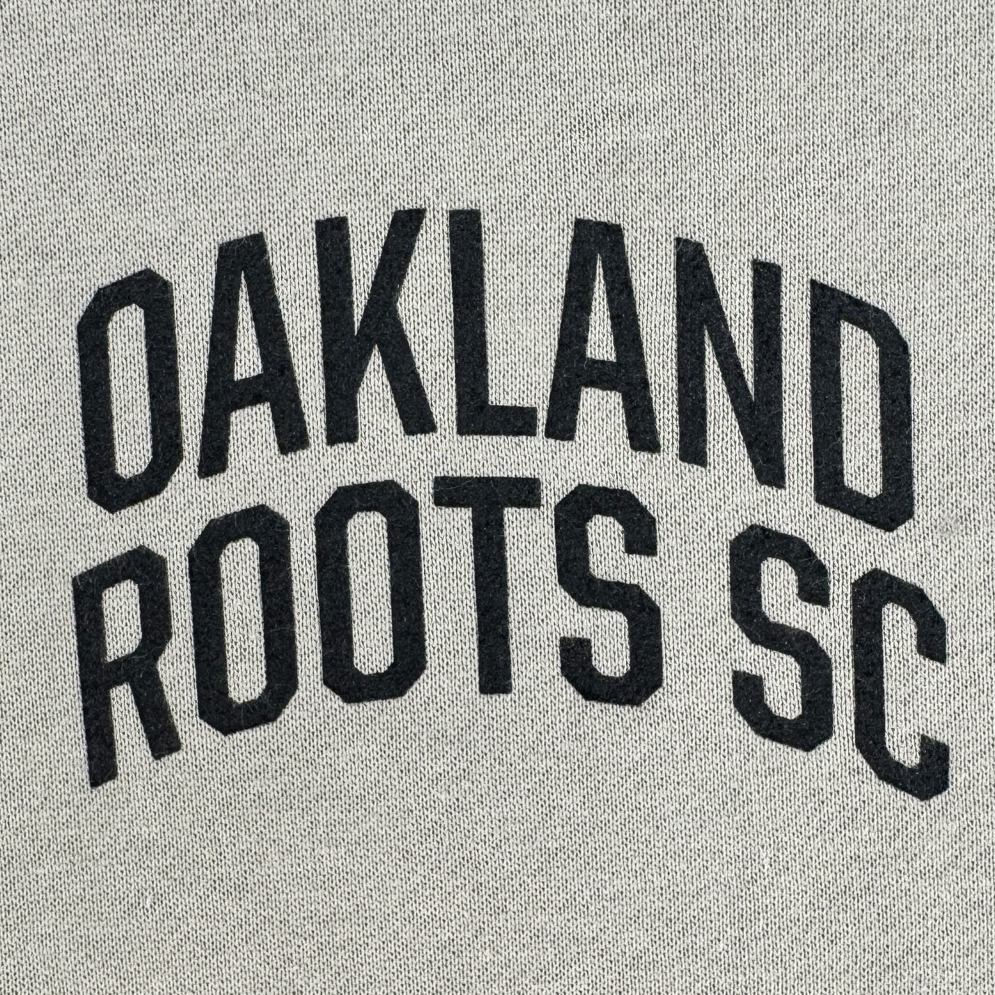 The image showcases "OAKLAND ROOTS SC" in bold, black, arched capitals on a light gray textured fabric, reflecting the style of the Oakland Roots SC Classic Hoodie.