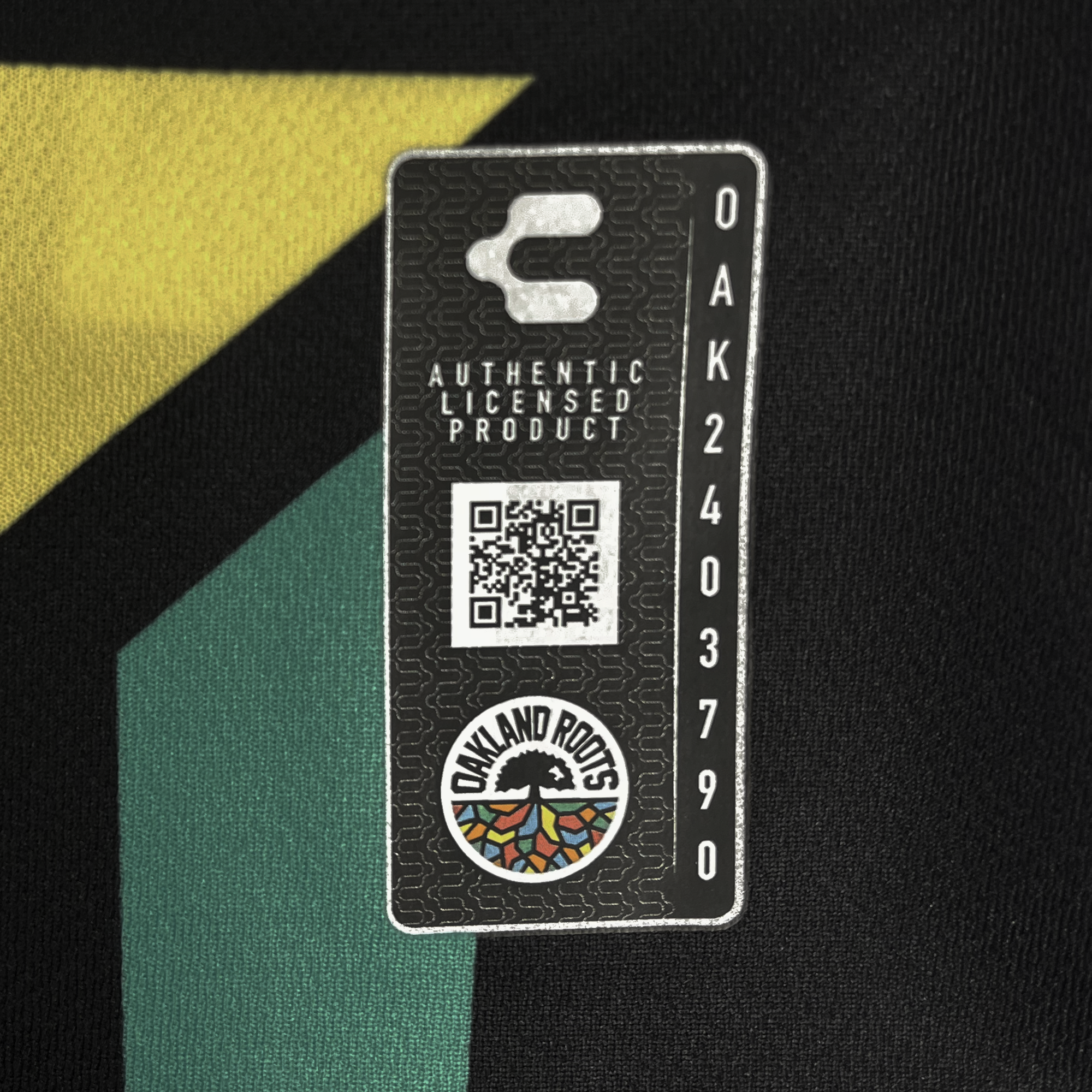 A close-up showcases a black fabric tag with a QR code, Oakland Roots logo with colorful roots on black, "AUTHENTIC LICENSED PRODUCT" text, side number "OAK2403790," and vibrant geometric shapes in the background, all part of the Unisex Oakland Roots SC 2025 First Kit ft. Anthem Blue Cross.