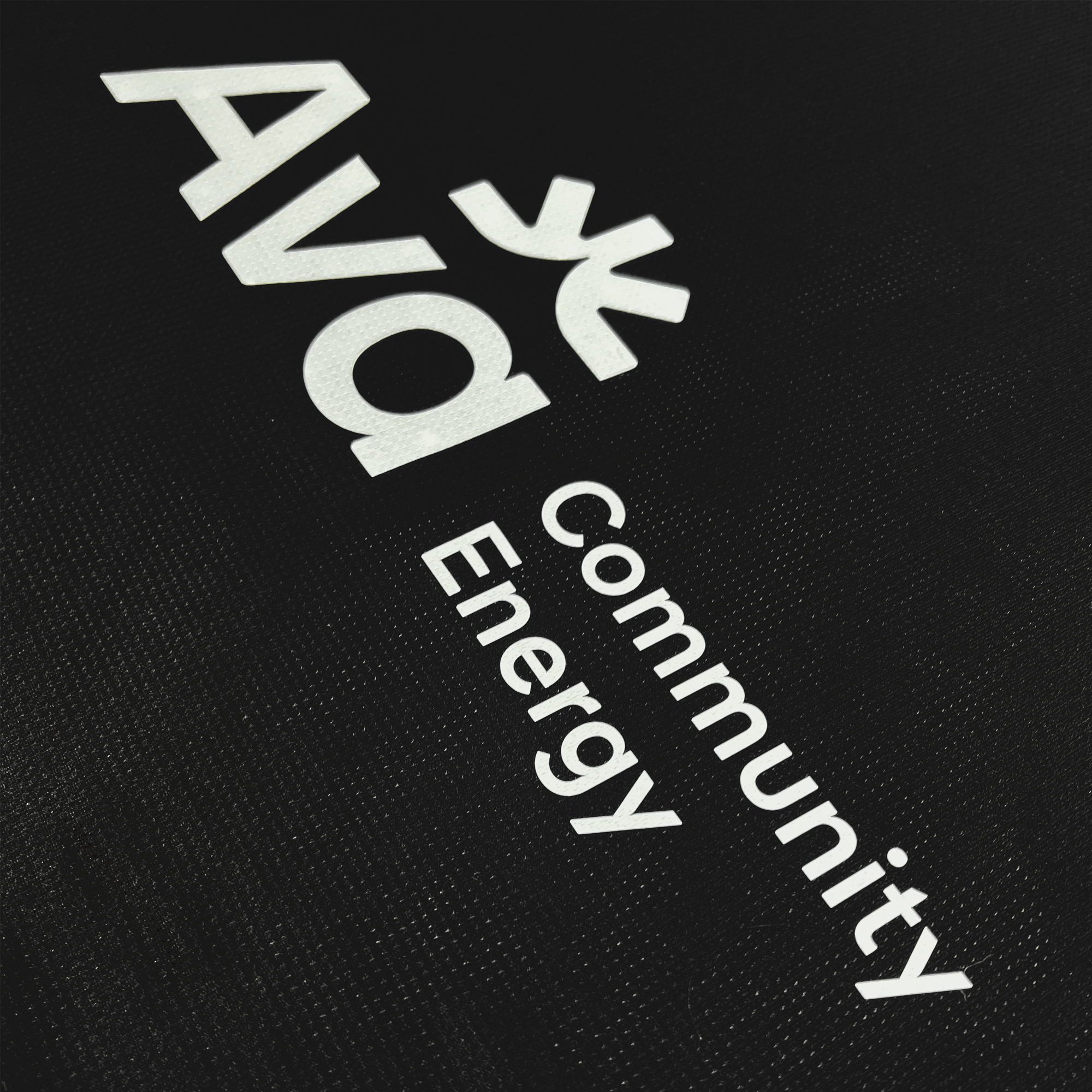 The unisex Oakland Roots SC 2025 First Kit features white text on a black base, reading "Ava Community Energy" in a modern font. The larger "Ava" has a leaf-like symbol above the "v," and the background's subtle texture resembles fabric or a matte surface.