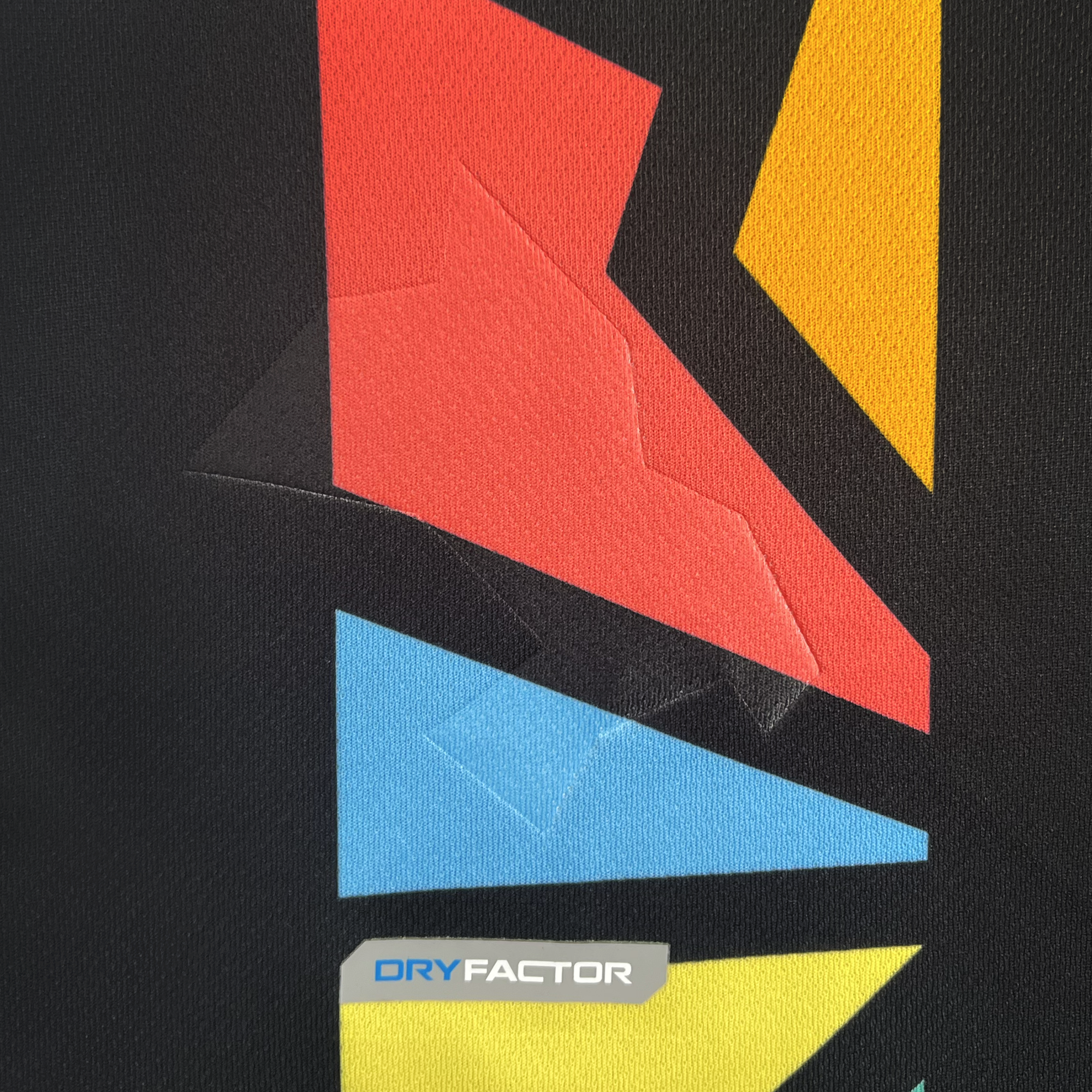 The detailed view of the Unisex Oakland Roots SC 2025 First Kit shows a fabric with geometric shapes in red, yellow, and blue on black. The "DRY FACTOR" logo appears at the bottom with "DRY" in blue and "FACTOR" in gray. Noticeable zigzag stitching adds texture to the surface.