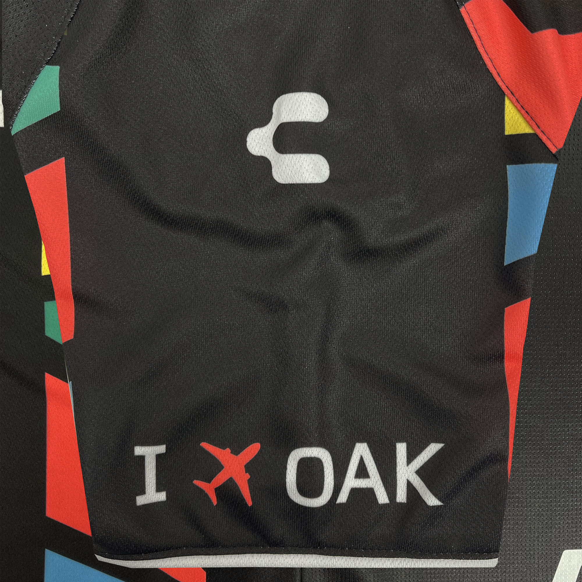 The Unisex Oakland Roots SC 2025 First Kit features a black fabric with a white "C" symbol, vibrant red, blue, yellow, and green geometric shapes for inclusivity, and a bold design completed by an "I" with a red airplane icon and "OAK" in white.