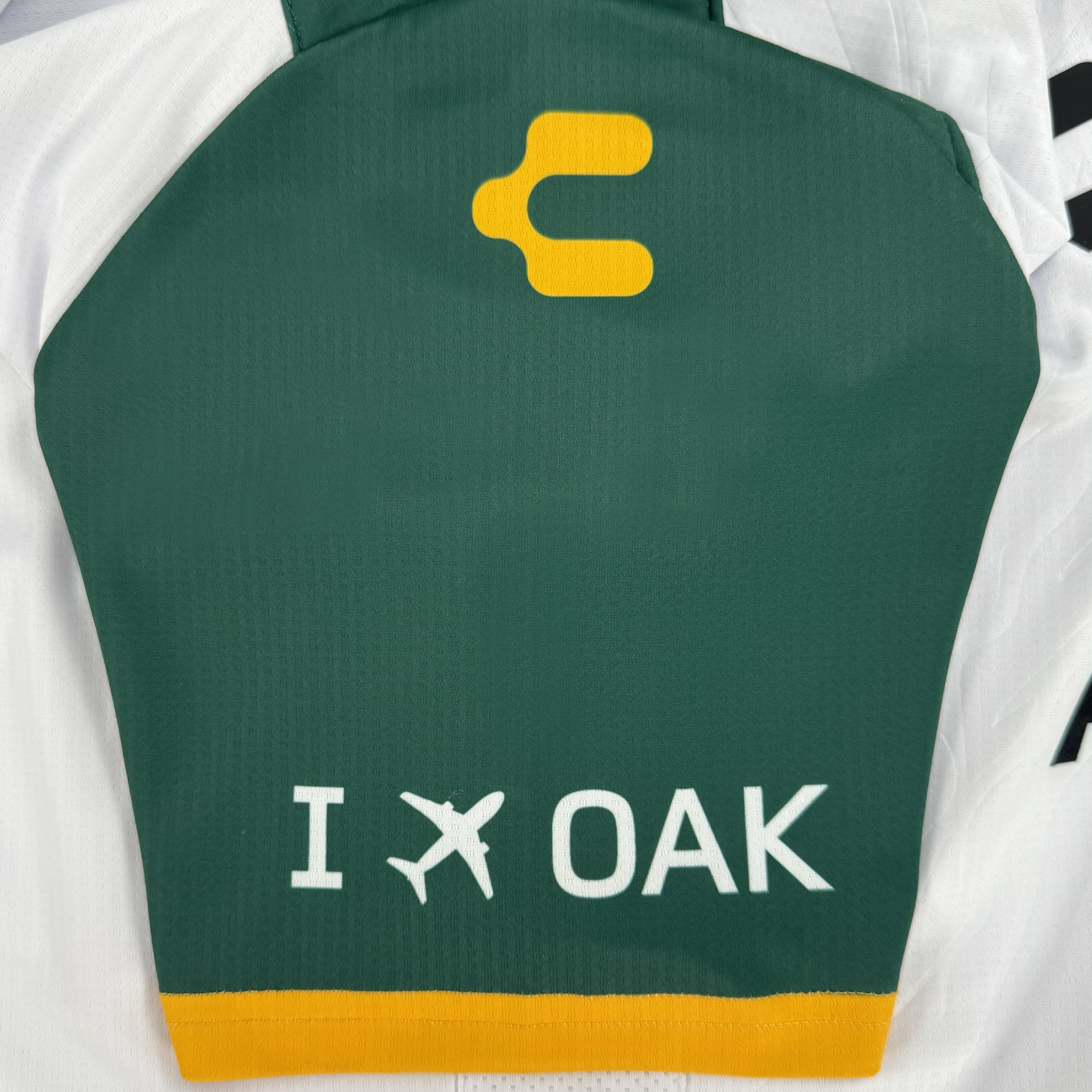 The Unisex Oakland Roots SC 2025 Second Kit by Oakland Roots SC features green and white fabric with a yellow emblem, celebrating Oakland sports. It reads "I [airplane symbol] OAK" and has a yellow-trimmed bottom edge.