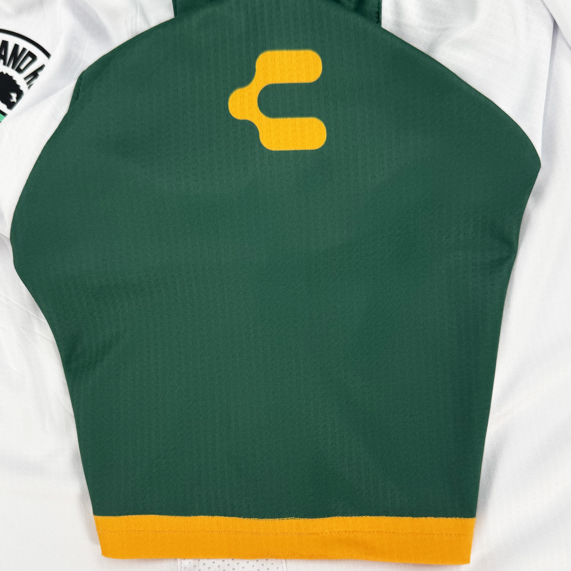 The Unisex Oakland Roots SC 2025 Second Kit features a green section with an orange abstract "C" or "E" logo, nodding to Oakland sports history. An orange stripe trims the lower edge. The white fabric includes a partially visible black and white patch on the left. Brand: Oakland Roots SC, Anthem Blue Cross sponsor.