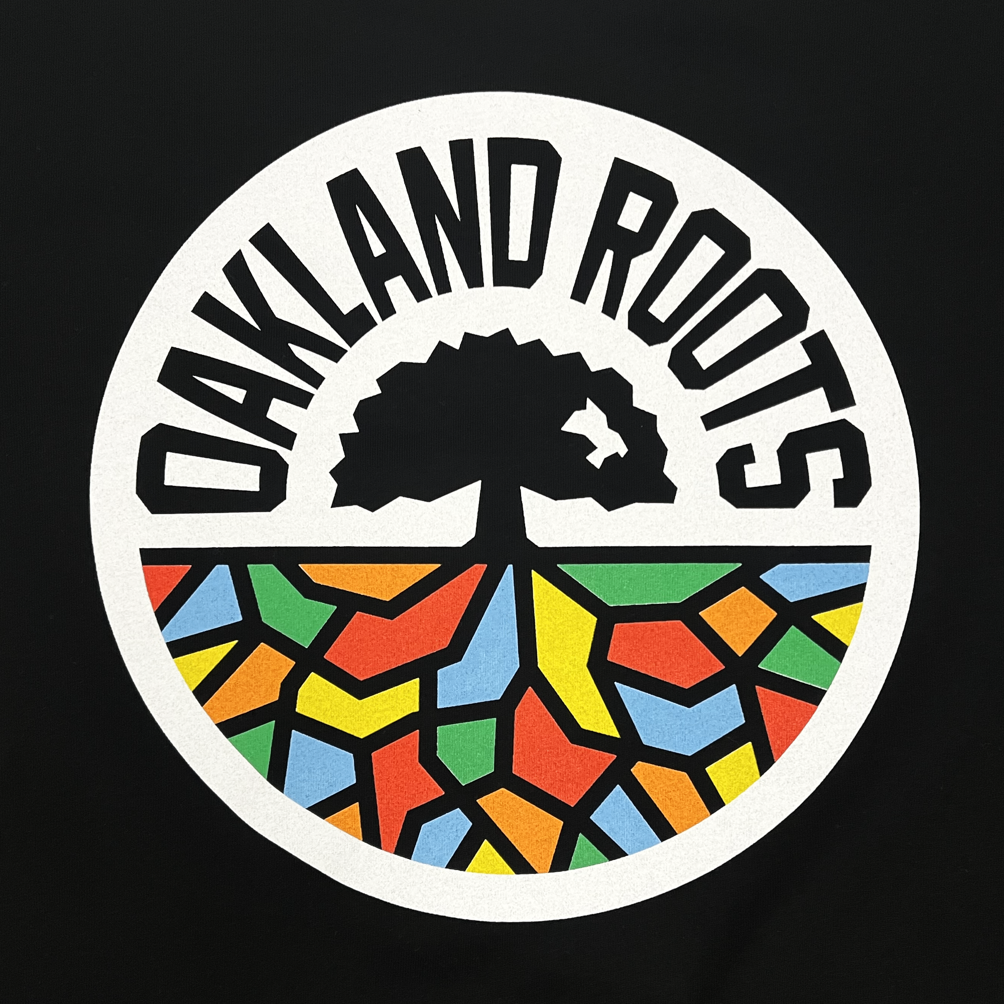 The Women's Oakland Roots SC Classic Relaxed V Neck Tee from Oakland Roots SC features a circular logo with a black tree silhouette and roots extending below. These roots intersect with a vibrant mosaic pattern comprised of shapes in red, yellow, green, and blue. The text "OAKLAND ROOTS" elegantly curves around the top half of the circle on a black background.