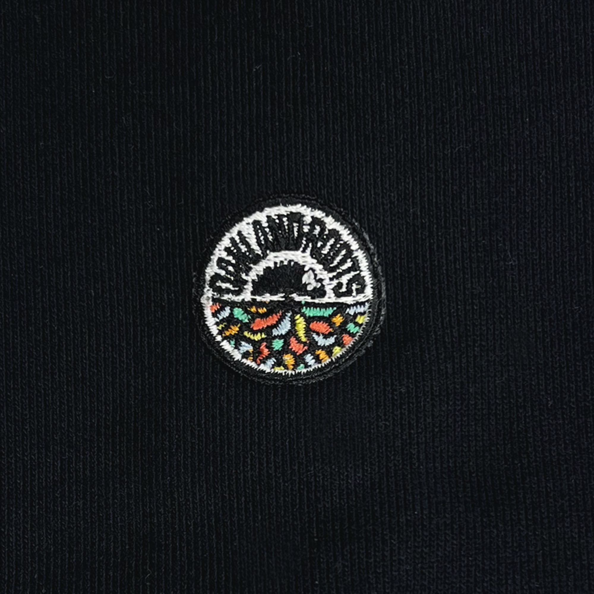 Close-up of a circular embroidered patch on black fabric. The patch features a mountain above a body of water with vibrant orange, green, and yellow shapes below. "CHAMUROS" arcs over the mountain in white text, reminiscent of the community spirit often seen in Oakland Roots SC's social good initiatives. This detailed design adds unique flair to the Members Only Polo by Oakland Roots SC.