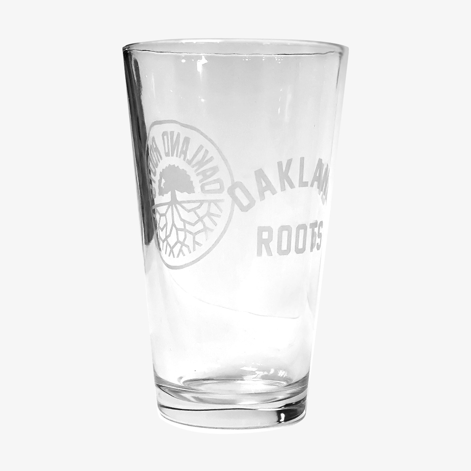 The Oakland Roots SC Pint Glass is a clear beer glass etched with the proud "Oakland Roots" logo, representing Oakland's vibrant sports club. The circular emblem features a tree with roots extending downwards, accompanied by the "Oakland Roots" text to the right. Simple and elegant, it stands on a flat surface against a plain white background.