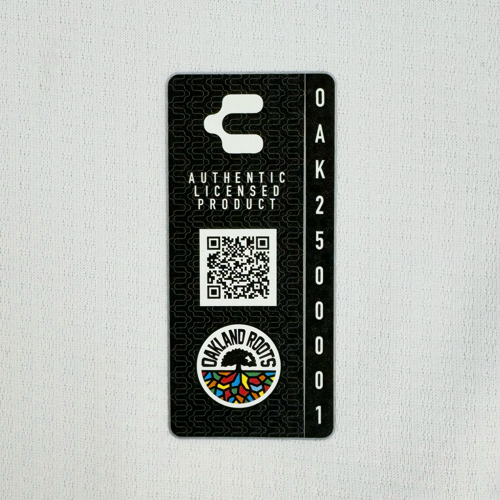 The black rectangular tag with a cut-out hook includes white text, "AUTHENTIC LICENSED PRODUCT" and "OAK2499999." It features a QR code and the "OAKLAND ROOTS" circular logo with a vibrant tree design, celebrating Oakland sports history. From the Unisex Oakland Roots SC 2025 Second Kit by Oakland Roots SC.