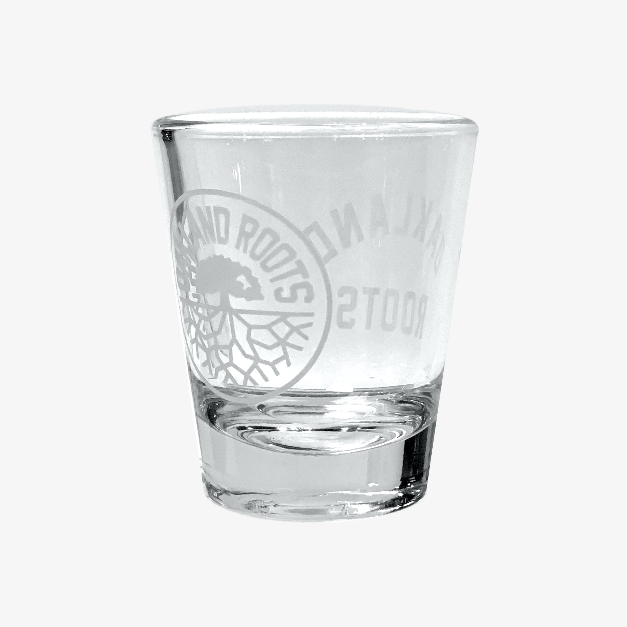 The Oakland Roots SC Shot Glass, branded by Oakland Roots SC, features a distinct circular logo showcasing a tree with roots and is prominently inscribed with "OAKLAND ROOTS Sports Club" on the front. The glass is displayed empty against a plain white background.