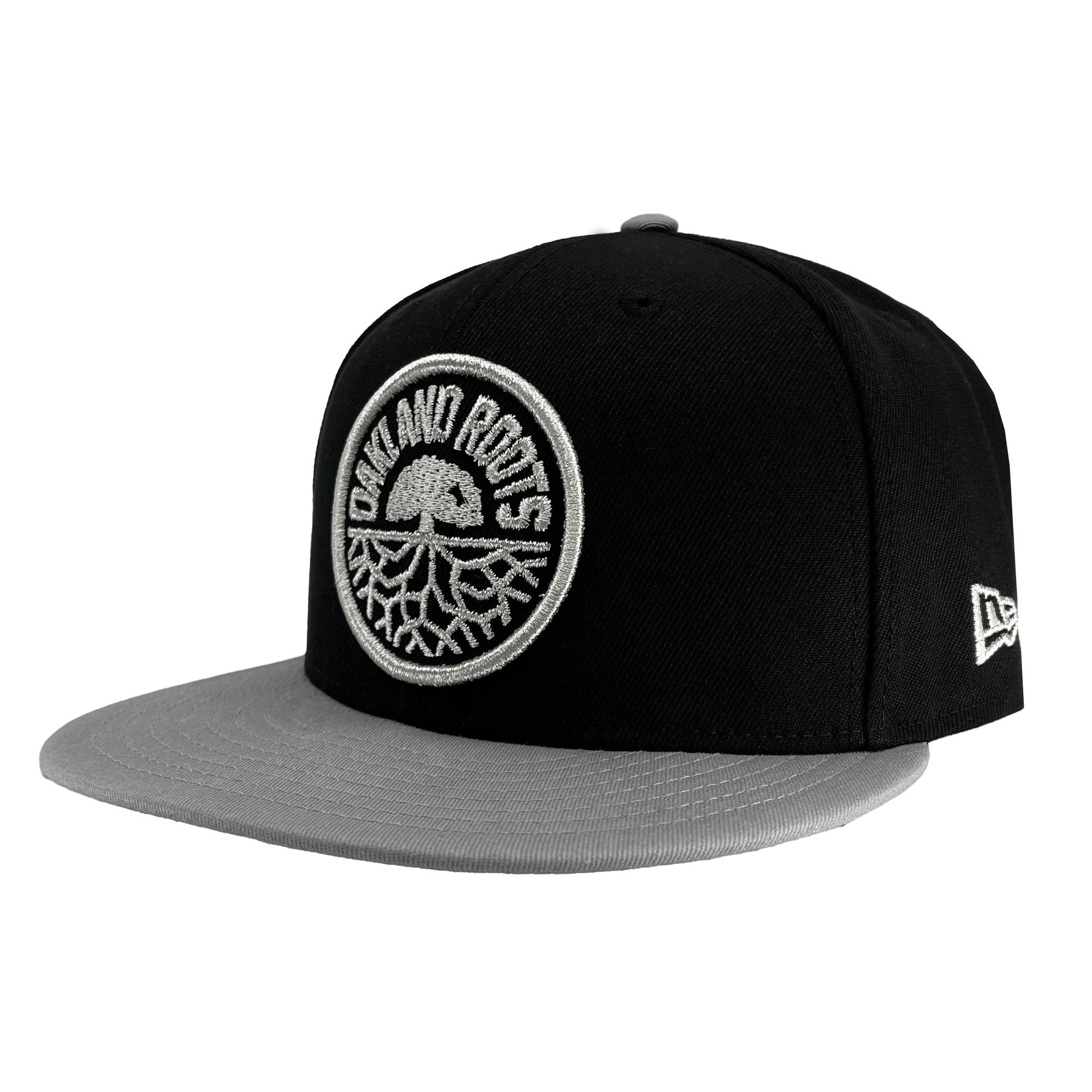 The New Era Oakland Roots SC 9FIFTY Cap, a black and gray snapback hat with a flat brim, features an embroidered circular logo on the front panel with the words "OAKLAND ROOTS" surrounding a stylized tree design. Its gray brim and small brand logo on a white patch enhance its sleek sports club style.