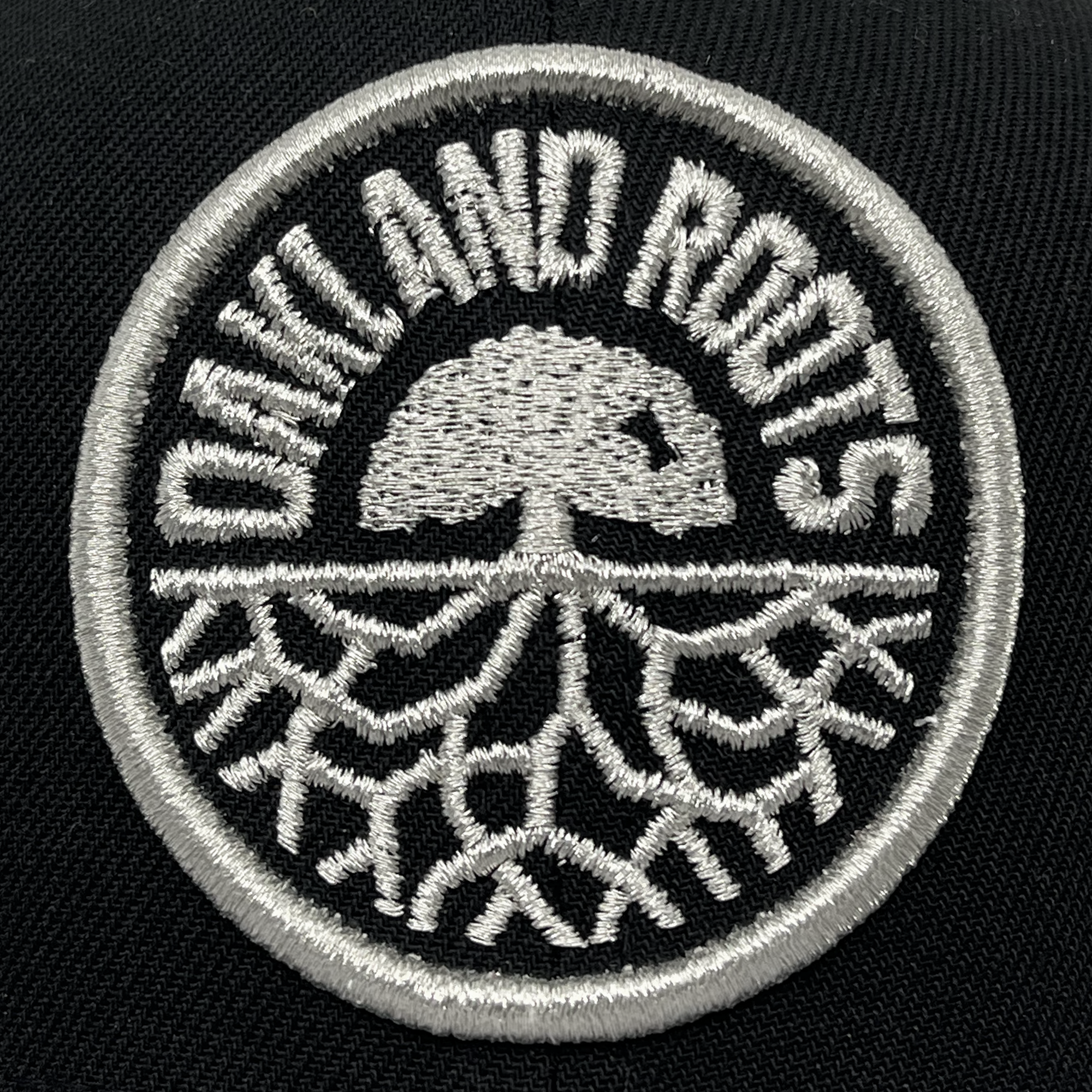 A circular embroidered patch shows the logo of the Oakland Roots SC. The patch is black with white embroidery, featuring a stylized tree with exposed roots. The words "OAKLAND ROOTS" curve around the top edge of the circle, making it a perfect addition to any New Era Oakland Roots SC 9FIFTY Cap by Oakland Roots SC.