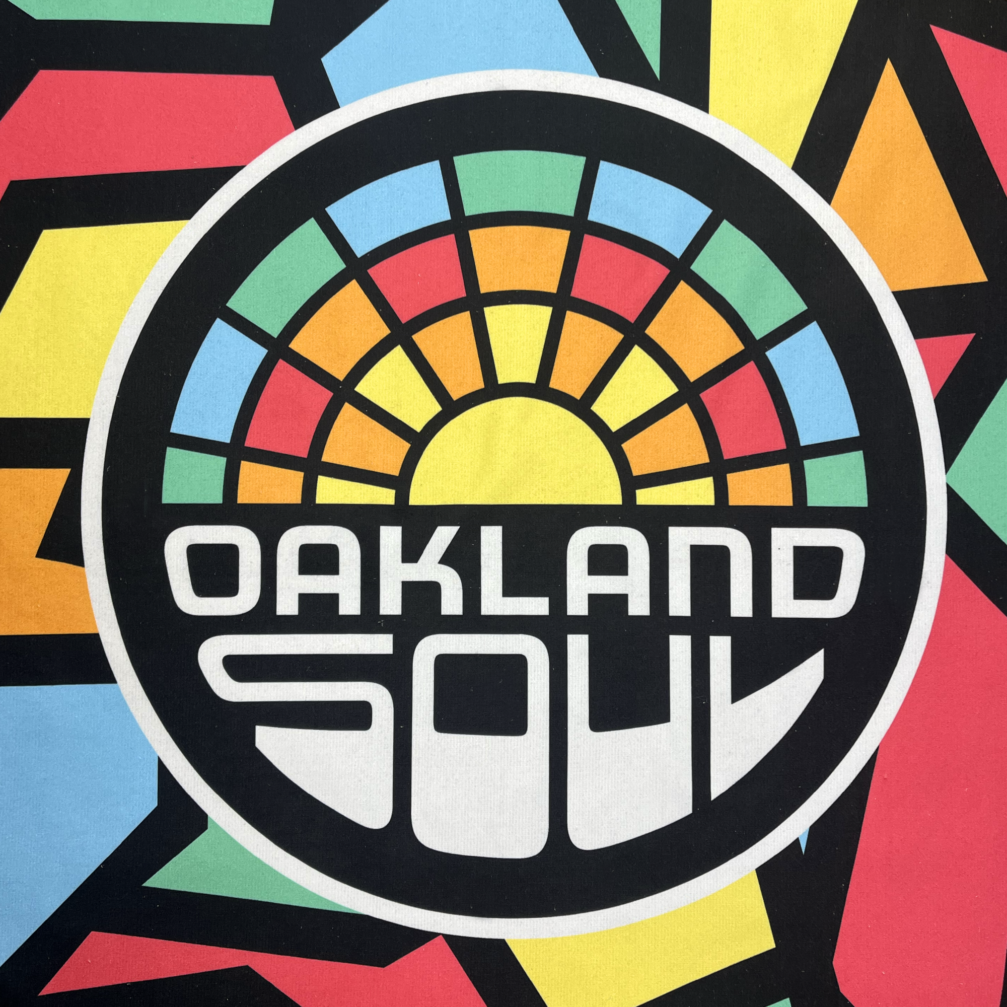 Introducing the Oakland Roots SC & Soul Towel by Oakland Roots SC: a dynamic and vibrant accessory featuring a bold logo inspired by the Oakland Roots Sports Club. The design showcases the text “OAKLAND SOUL” beneath a stylized sun, all enclosed within a circular frame. The background boasts an abstract geometric mosaic in green, red, blue, yellow, and black. This modern design captures dynamic energy and promotes social good vibes.