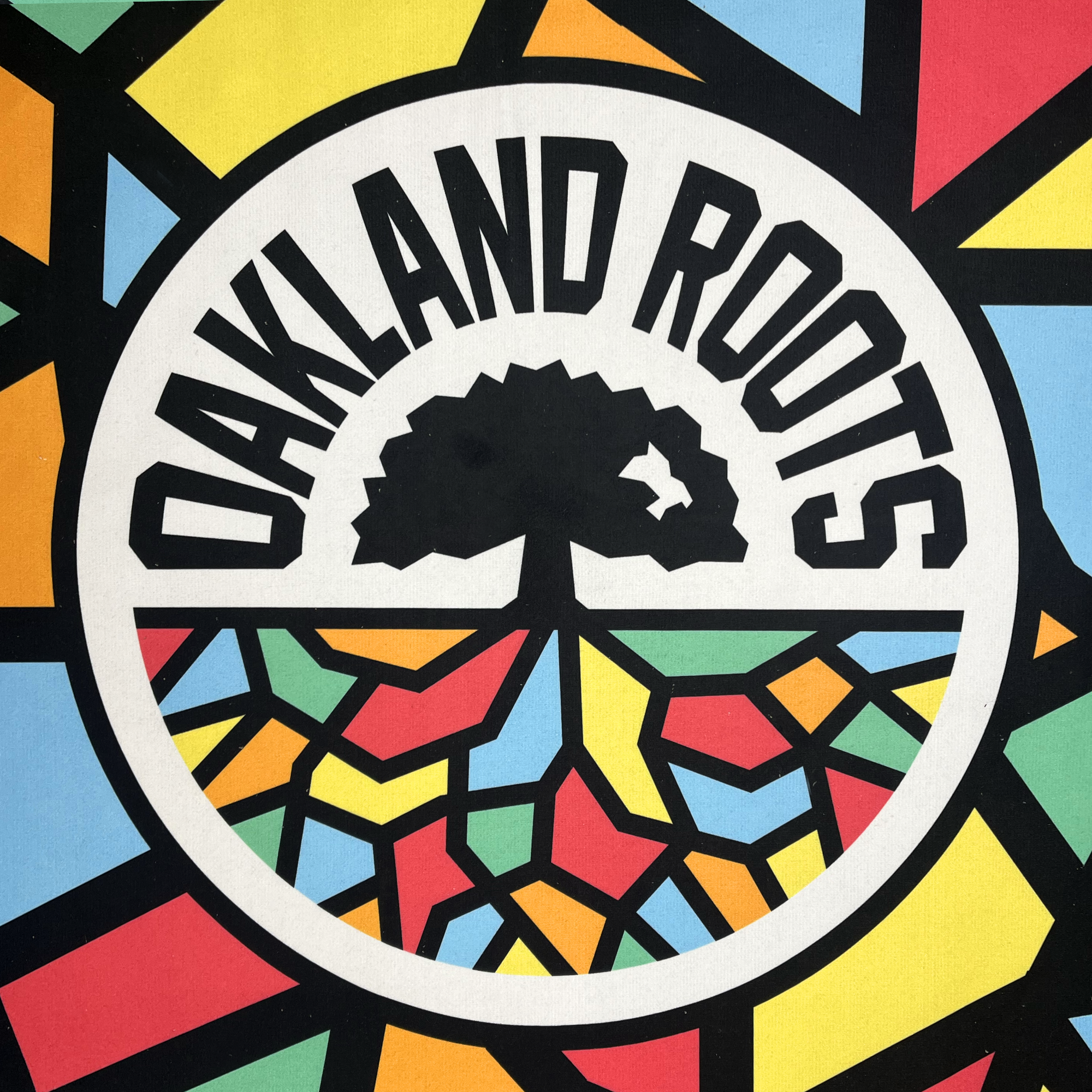 The Oakland Roots SC & Soul Towel by Oakland Roots SC showcases a round logo with a stained-glass design, prominently featuring "Oakland Roots" in large, bold letters. Within the circle, a tree silhouette stands above colorful shapes that resemble roots or a mosaic pattern. This vibrant emblem symbolizes the Oakland Roots Sports Club and their commitment to social good.