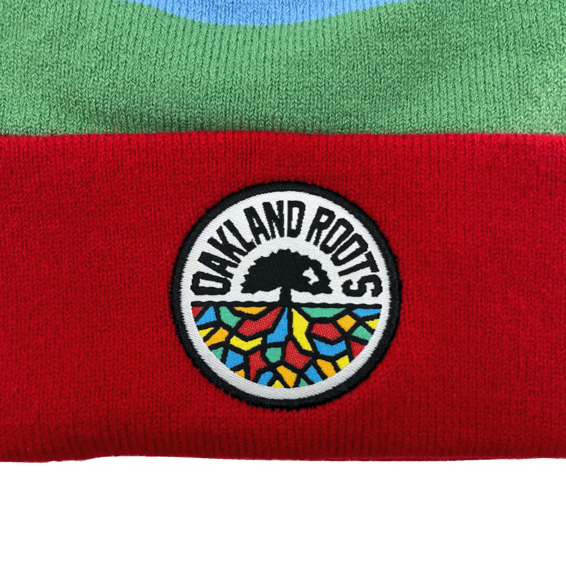 A close-up of the Oakland Roots SC Mosaic Stripe Beanie shows a textured fabric with green on top and red on the bottom. In the center, a circular patch features a stylized tree with roots spreading out, colored in vibrant, mosaic-like patterns. The words "OAKLAND ROOTS Sports Club" are displayed in a semicircle above the tree.