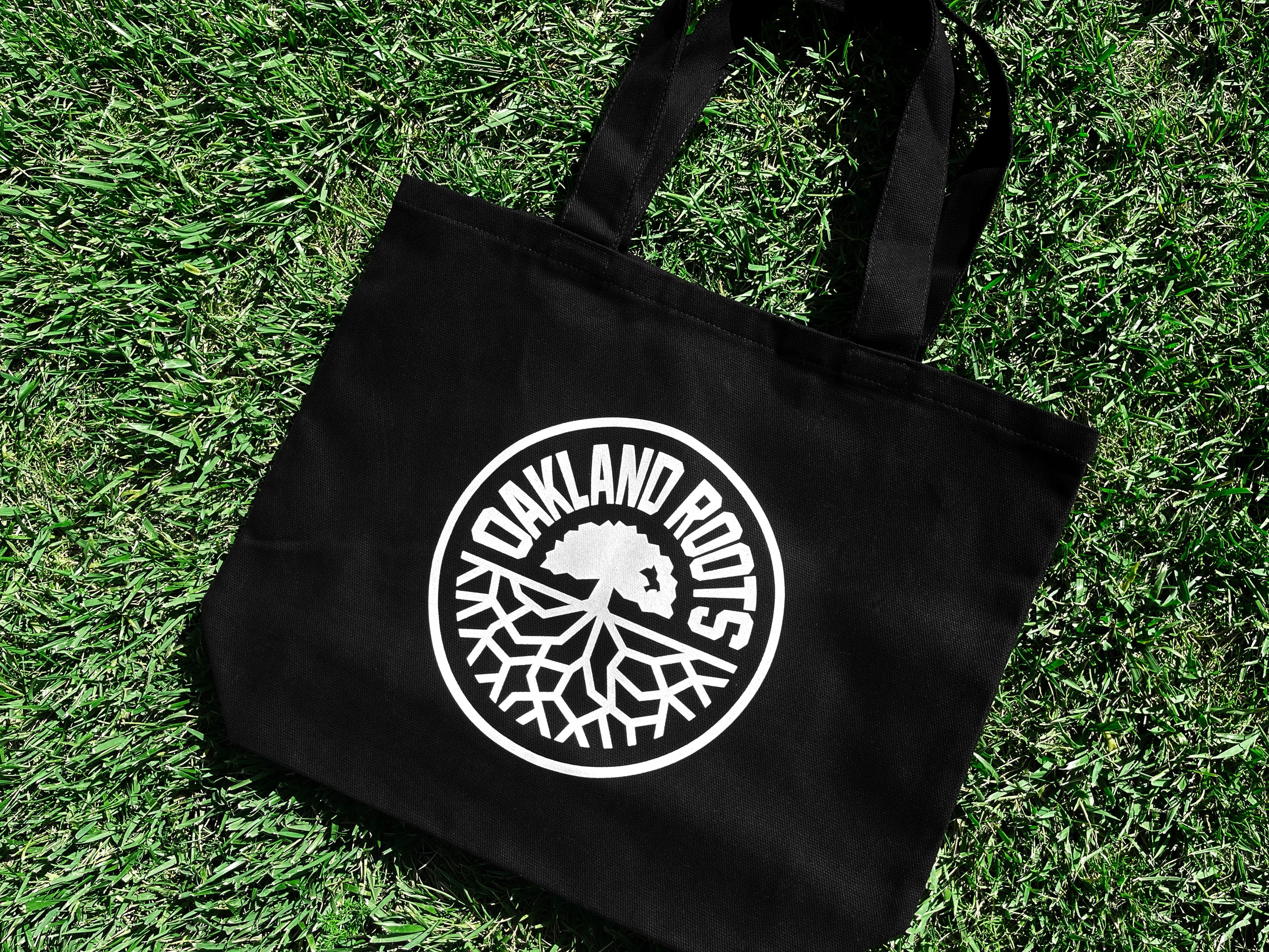 An Oakland Roots SC Tote from Oakland Roots SC is lying on a grassy surface. The black bag features a white circular logo in the center with the text "Oakland Roots Sports Club" surrounding an oak tree and interwoven roots graphic, reminiscent of the iconic Oaklandish design.