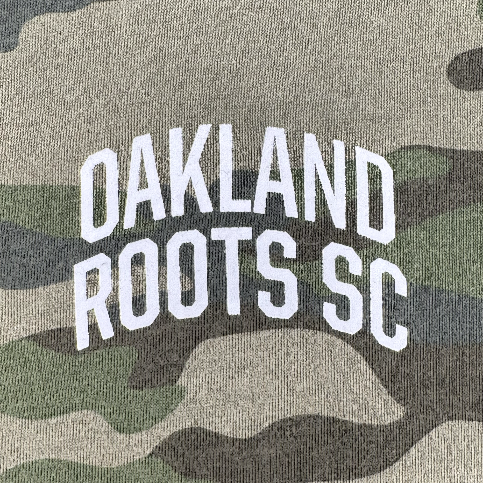 The image features the "OAKLAND ROOTS SC" text in bold, white uppercase letters on a fabric with a unique camouflage design, reflecting the vibrant spirit of Oakland Roots SC. Resembling an Oaklandish hoodie, it blends shades of green, brown, and beige—classic for the Oakland Roots SC Classic Zip Hoodie.