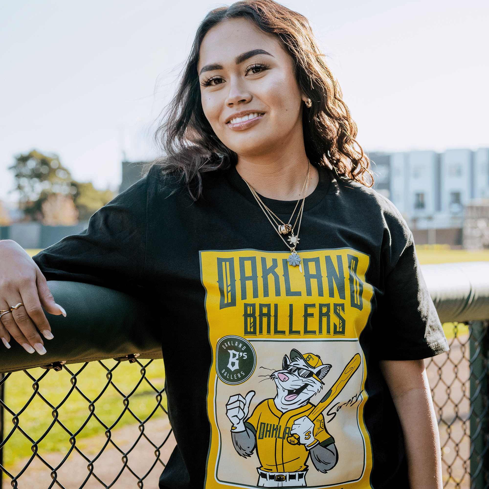 A woman with long curly hair stands by a fence wearing the Oakland Ballers Scrappy Rookie Card Tee, featuring mascot Scrappy the Rally Possum in a baseball uniform. She smiles slightly, with blurred trees and buildings in the background under a clear sky.