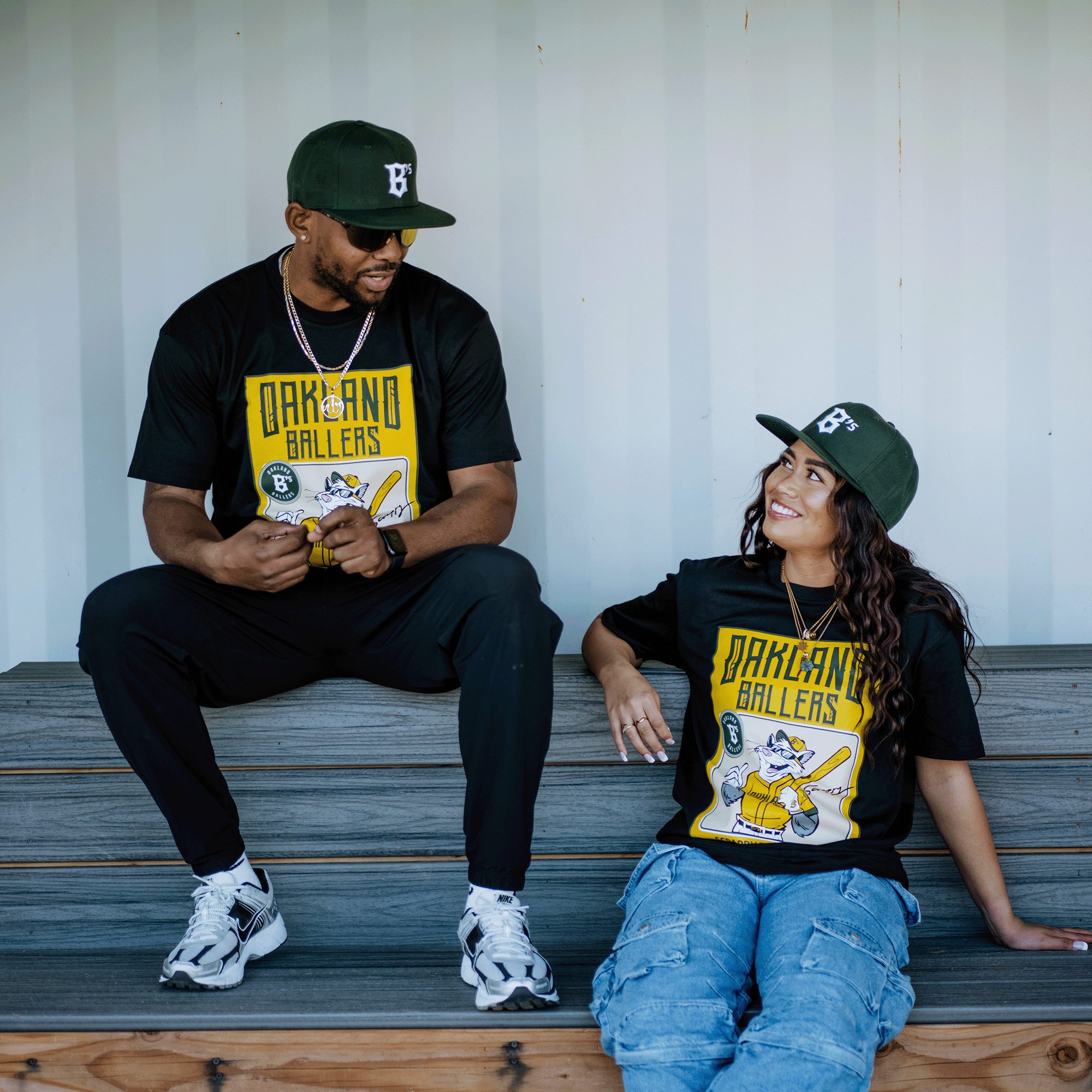 Two people in matching "Scrappy Rookie Card Tee" by Oakland Ballers and green caps with Scrappy the Rally Possum sit on wooden steps. The man, in black pants and white sneakers, chats with the woman in blue jeans and white sneakers. Both smile against a crisp white background.