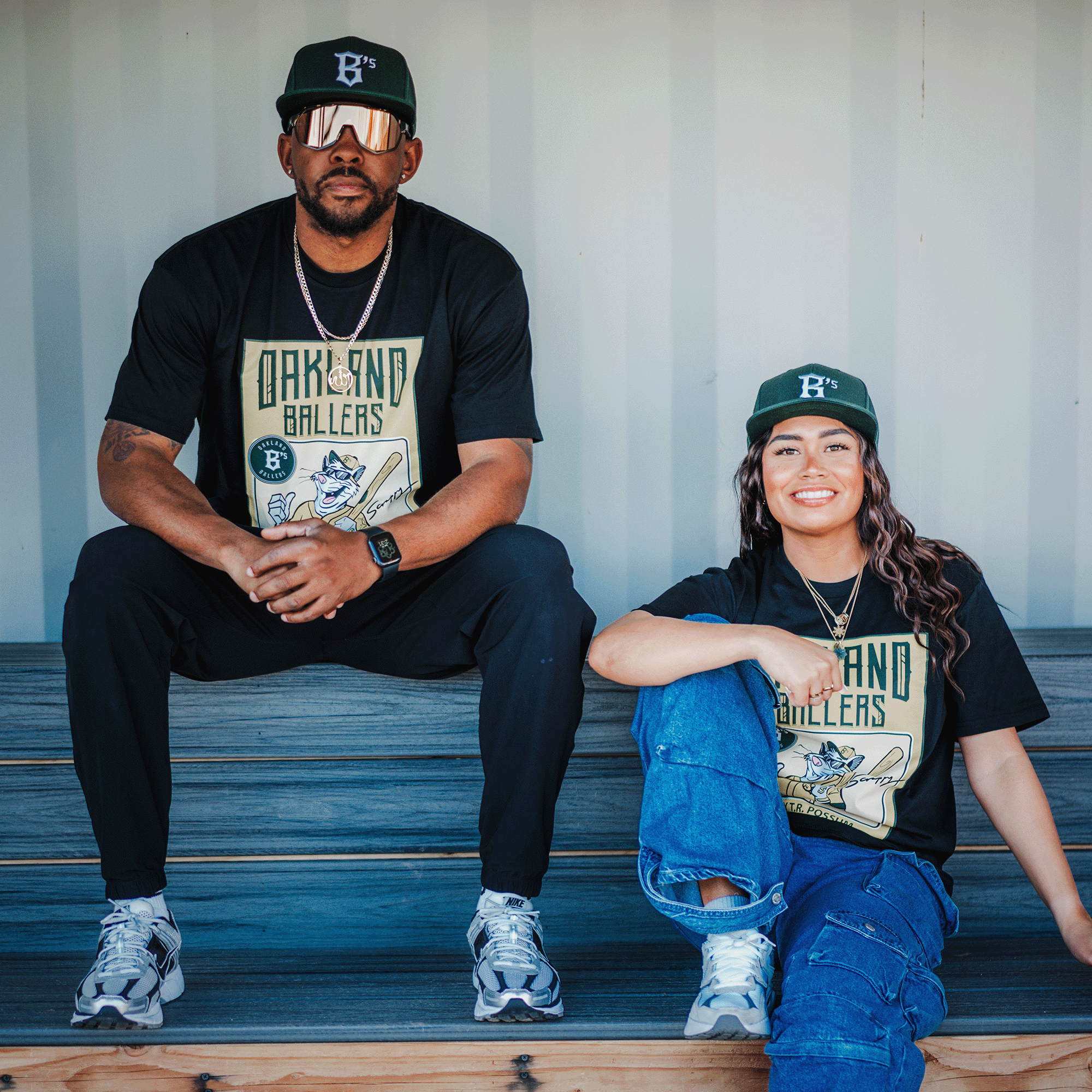 Two people in black "Scrappy Rookie Card Tee" by Oakland Ballers and green caps sit on wooden steps against a gray wall. The man wears sunglasses and black pants, while the woman in ripped jeans sits with one knee up, both exuding a relaxed vibe reminiscent of the Rally Possum spirit.