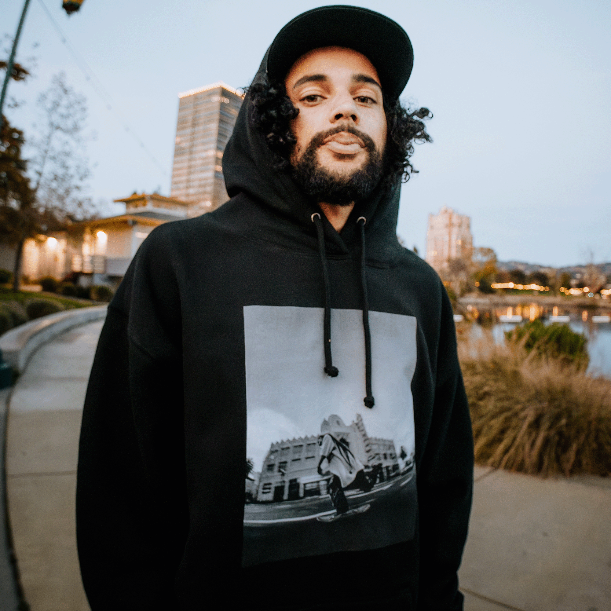 A person with curly hair and a beard, resembling a skateboarder, is sporting the "Skateboarding is Beautiful Hoodie" by Oaklandish in black, along with a black cap. They are outdoors in an Oakland park setting, surrounded by buildings near a paved path and body of water during the evening.