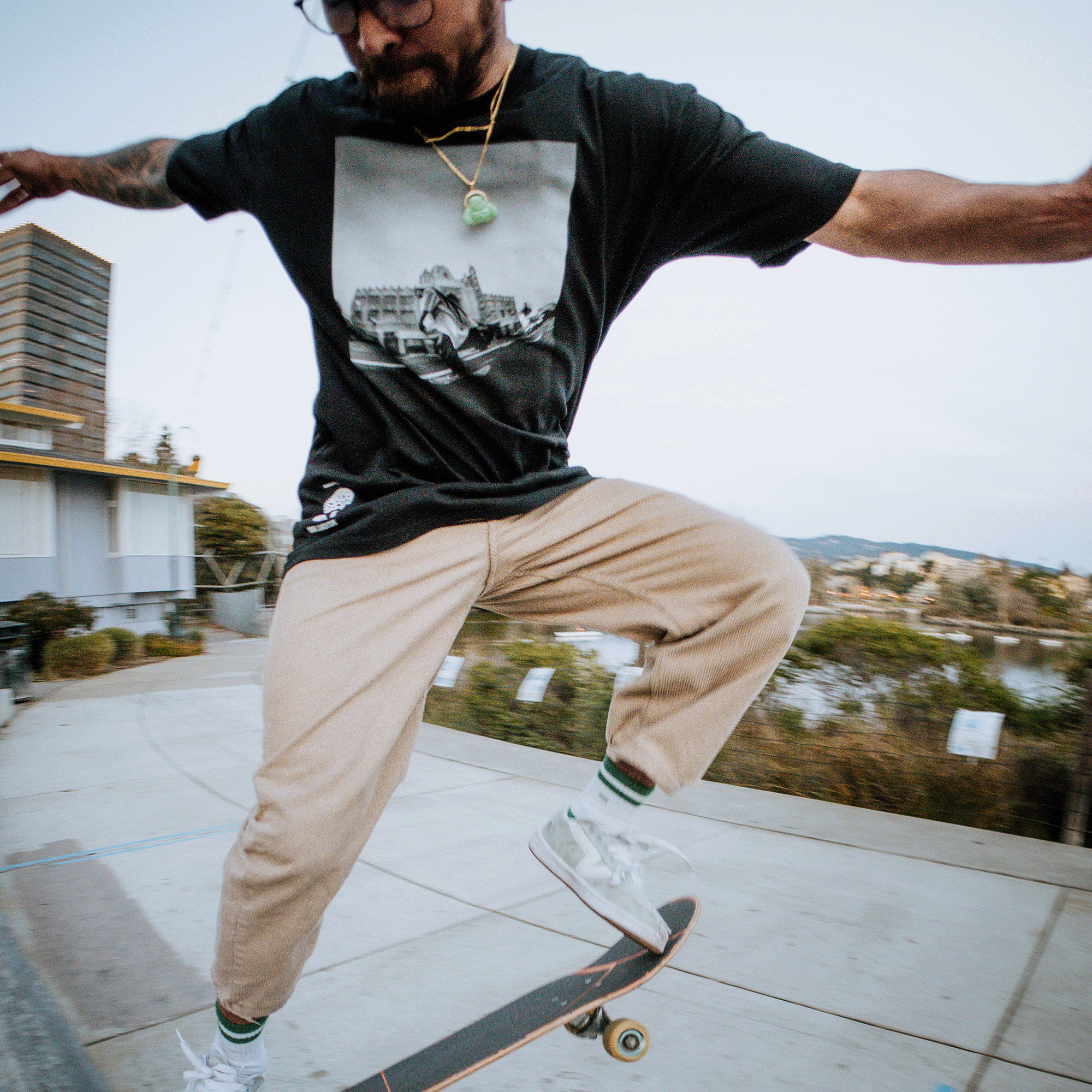 A person wearing the Oaklandish "Skateboarding is Beautiful Tee" and beige pants is performing a skateboard trick mid-air in Oakland. Sporting glasses, a beard, and necklaces, they are captured in a dynamic and energetic scene through vibrant photography, set against an industrial area with buildings and trees on a cloudy day.