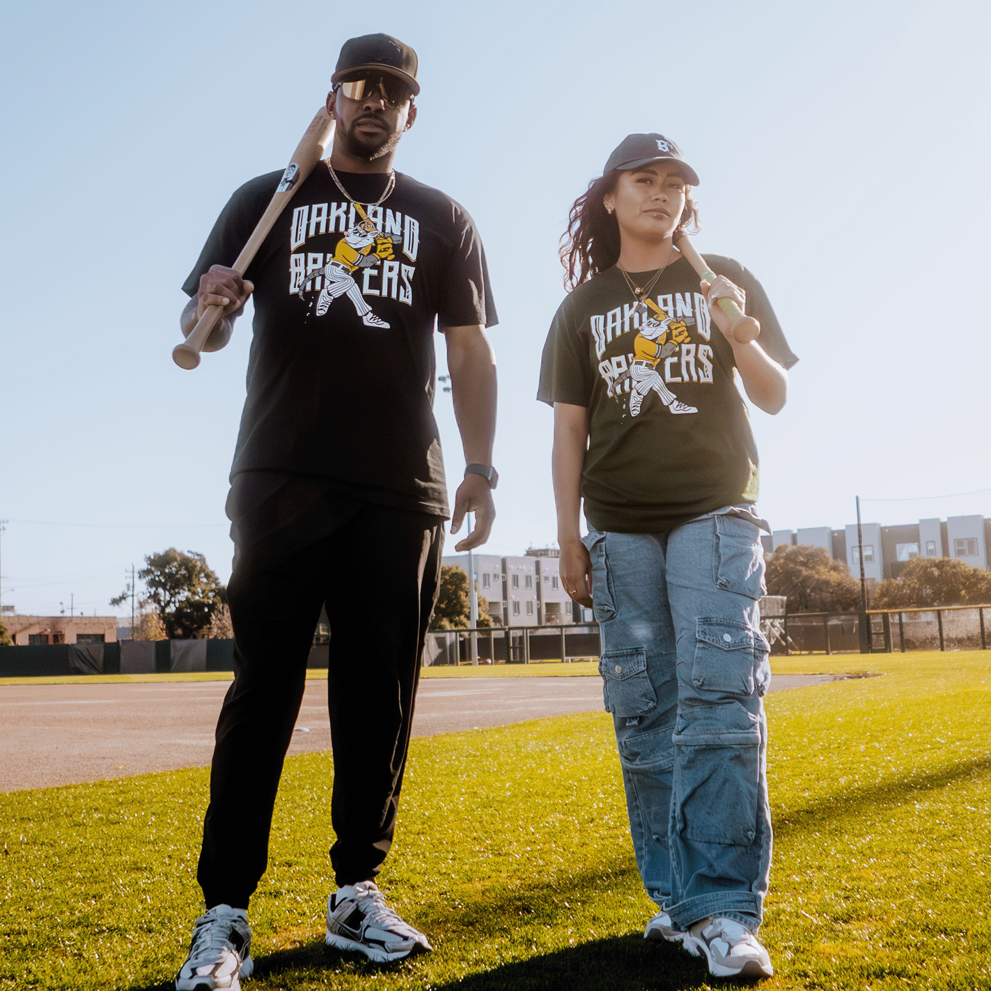 Two people stand on a sunny baseball field at Raimondi Park, proudly wearing "Oakland Ballers" Slugging Scrappy Tees with a yellow figure. The left holds a bat over their shoulder, while the right, in cargo jeans, rests theirs on the ground. Trees and buildings form the backdrop.