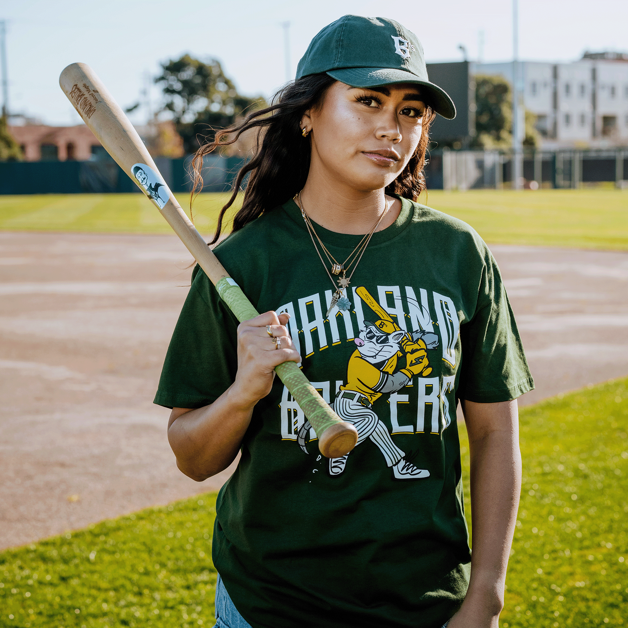 At Raimondi Park, a person stands on the baseball field, bat over shoulder. They wear an Oakland Ballers Slugging Scrappy Tee, matching cap, and jeans. The sun shines on the field, highlighting trees and buildings under a clear sky.