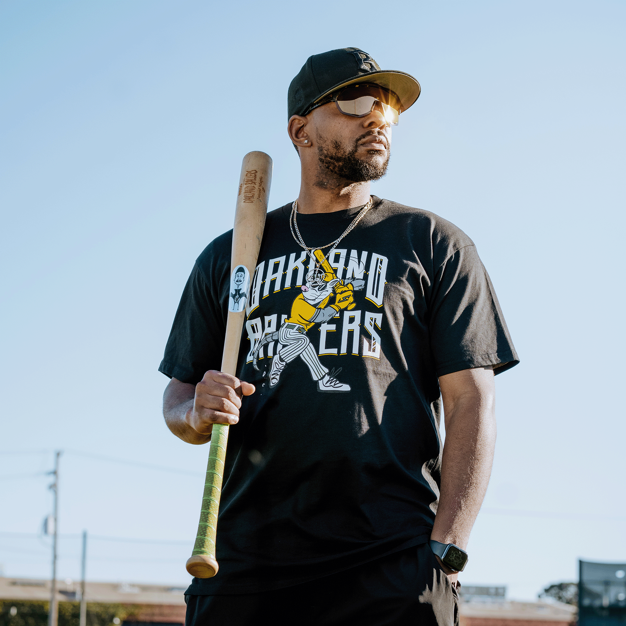 A man confidently holds a baseball bat over his shoulder, embodying the Oakland Ballers' spirit. He wears a black cap, sunglasses, and a "Slugging Scrappy Tee" featuring a baseball player illustration, posing against a clear blue sky and rooflines.