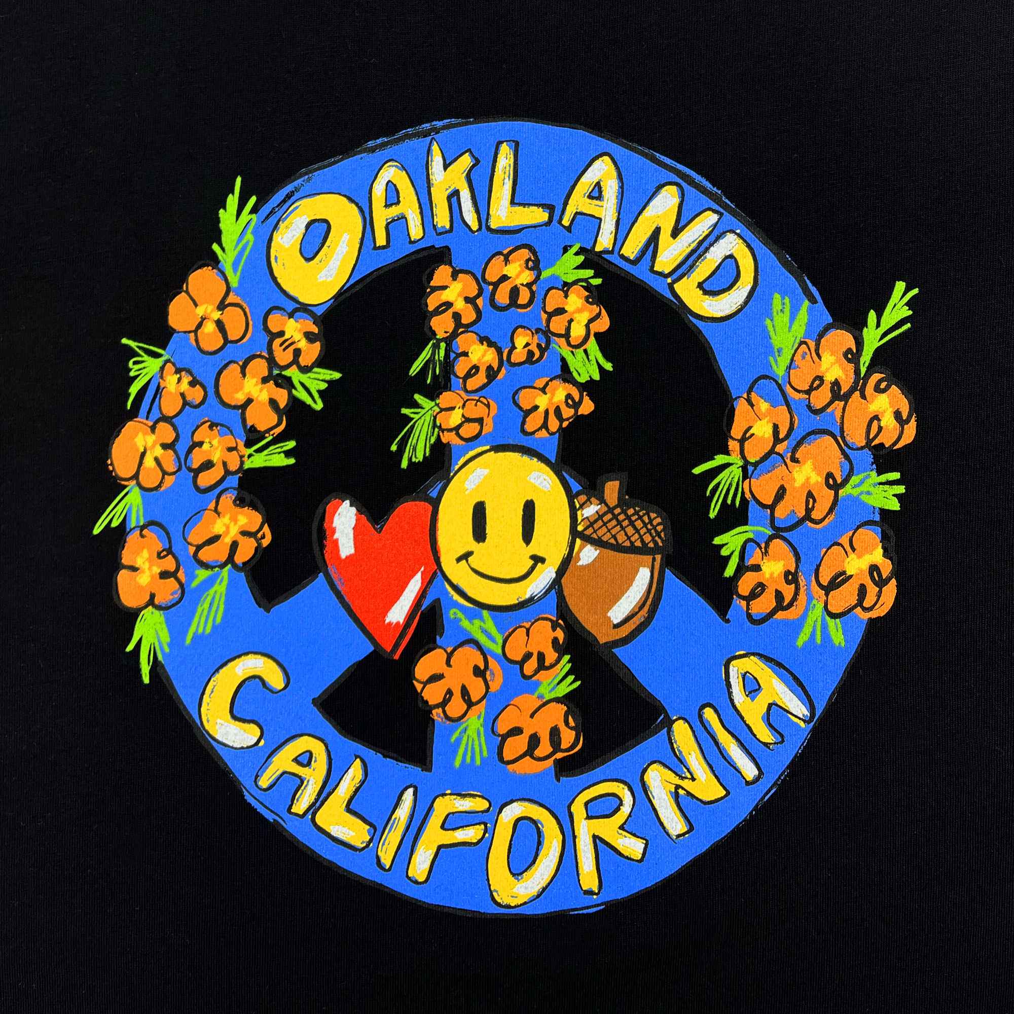 The Women's Smile Crop Tee by Oaklandish features a vibrant illustration on a black background. At the top, the word "Oakland" is displayed, while "California" sits at the bottom of a blue circular peace symbol. The design is decorated with orange flowers, a yellow smiley face, a red heart, and an acorn. This 100% cotton cropped t-shirt perfectly blends colorful and playful elements in women's sizing.