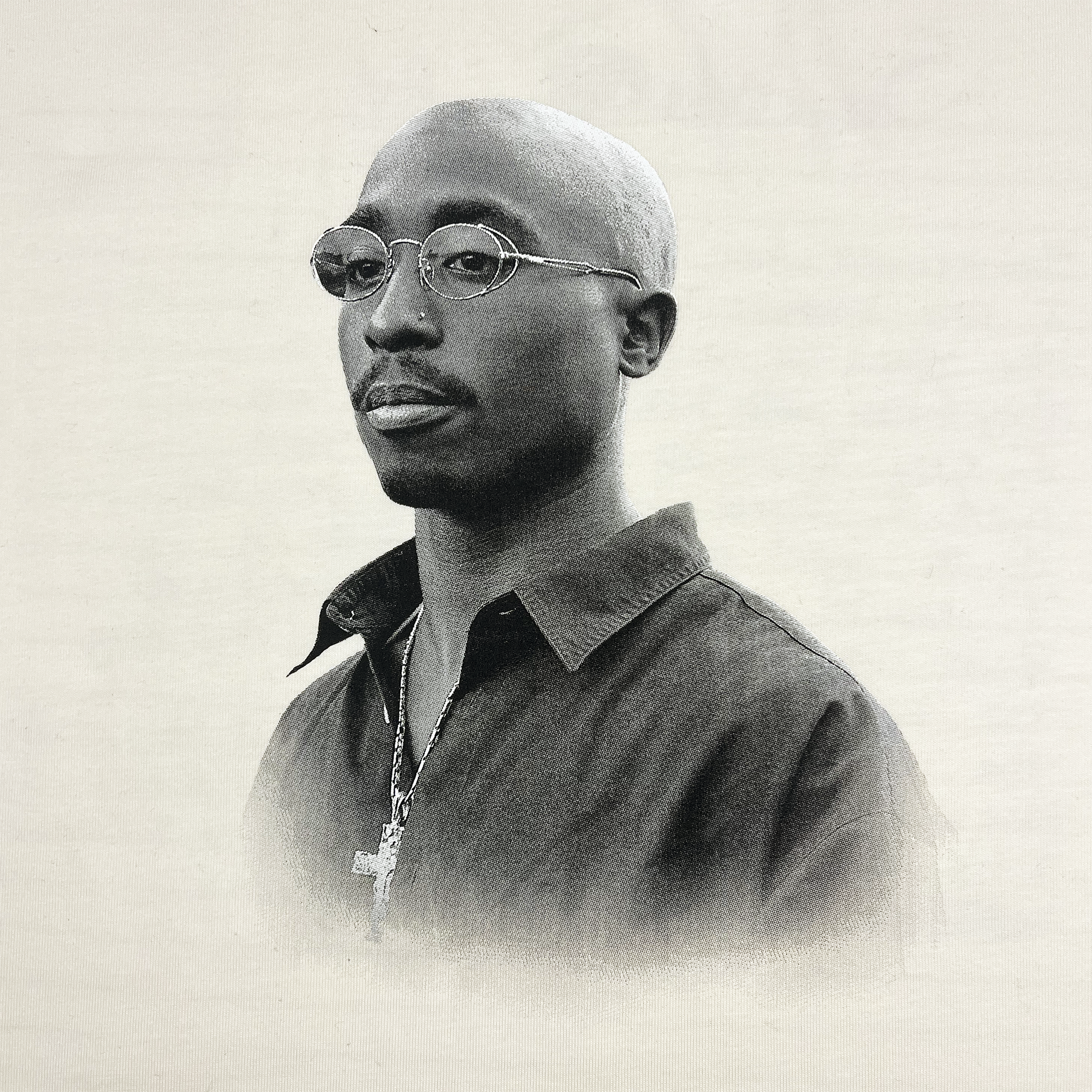 A black and white portrait captures a man in the Oaklandish Sometimes Eye Cry Tee, accessorized with glasses, a hoop earring, and a cross pendant necklace. His collared shirt complements the plain background that highlights his calm expression, reminiscent of hip-hop legends like Tupac Shakur.