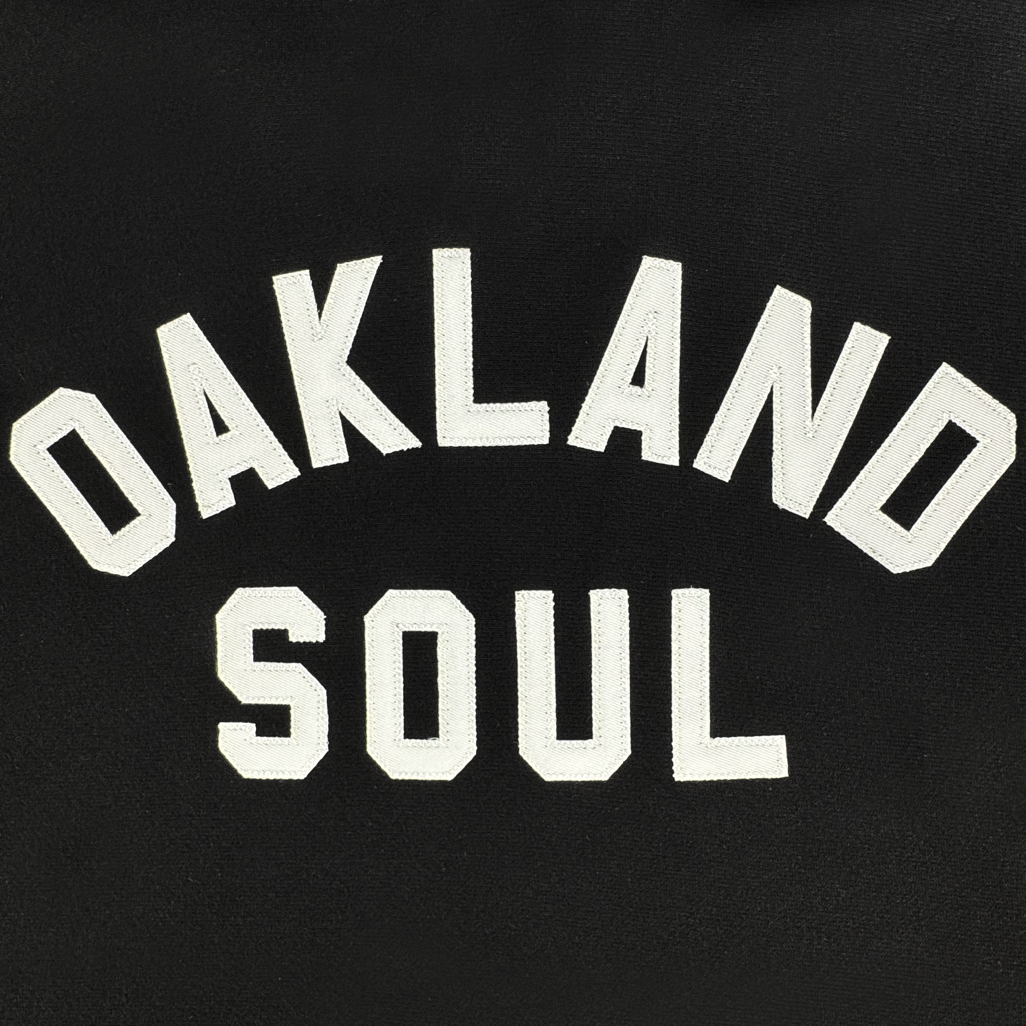 The Champion X Oakland Soul Wordmark Hoodie features a black backdrop with bold embroidered white letters. "OAKLAND" is arched above, and "SOUL" rests below in straight, blocky capitals, reflecting the vibrant spirit of newcomers Oakland Soul in the USL W-League of women's soccer.