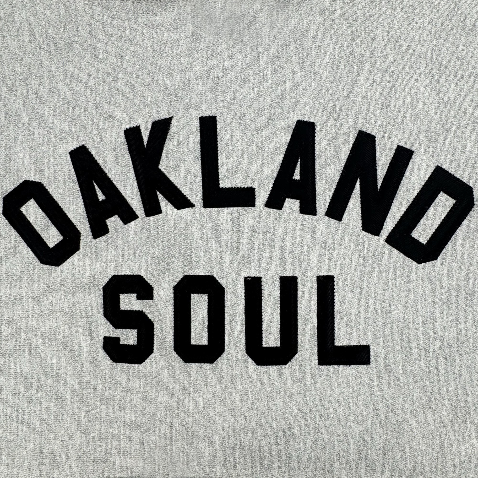 The Champion X Oakland Soul Wordmark Hoodie has a gray fabric background with bold black "OAKLAND SOUL" text in collegiate font, representing the spirited energy and passion of the USL W-League women's soccer team.