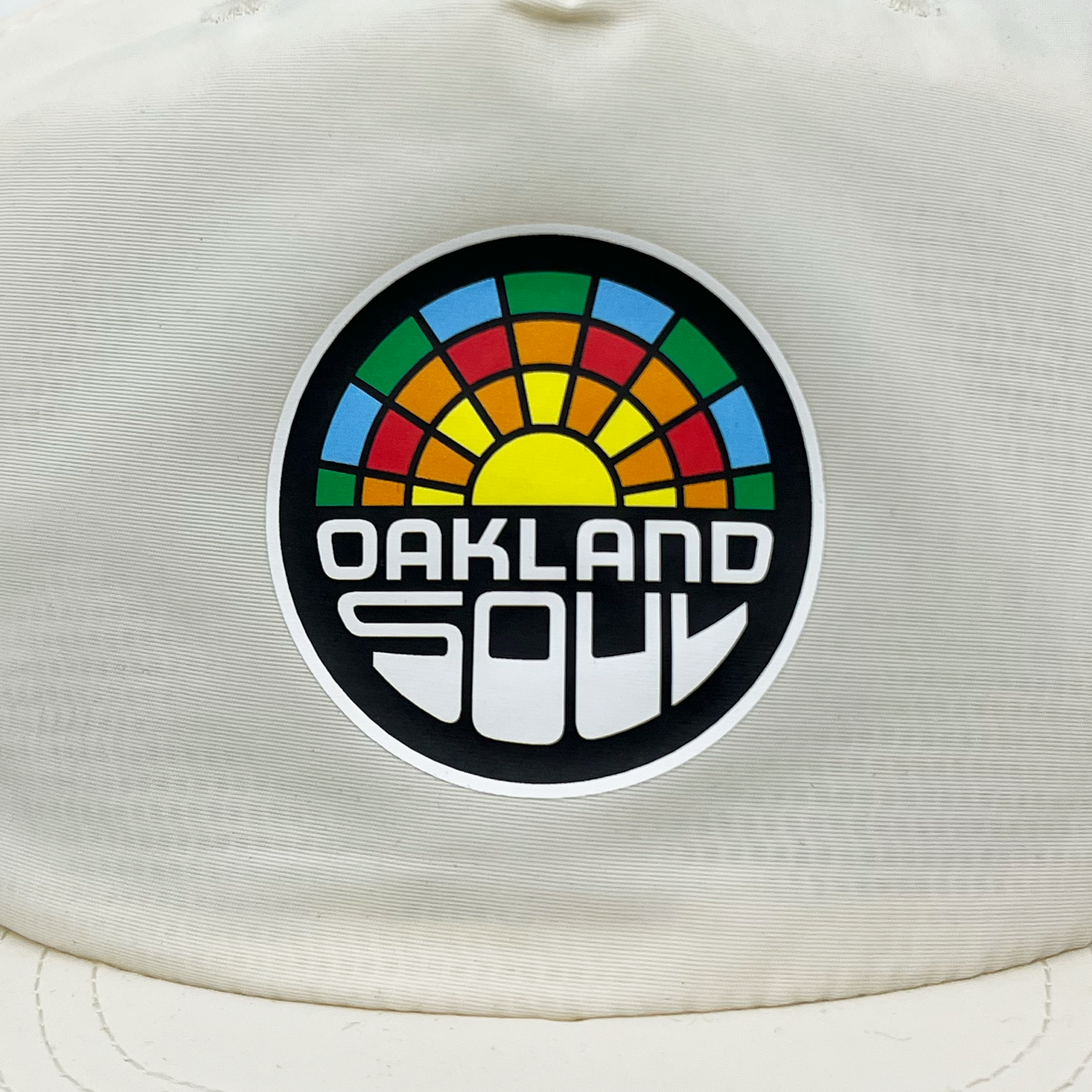 The Oakland Soul Surf Cap is a stylish white hat featuring a circular logo with a yellow sun at its center, surrounded by blue, green, orange, and red hues that create an eye-catching stained glass effect. Beneath this vibrant design, "OAKLAND SOUL" is boldly printed in modern white letters against a black background. Show your support for the Oakland Soul USL W-League women's soccer team in style with this fashionable accessory.