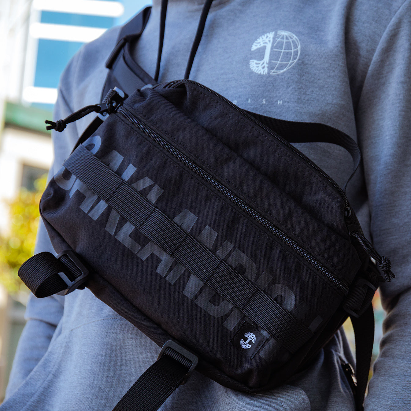  Close-up of the a man’s chest wearing a cross body black nylon hip bag with a Oaklandish wordmark and tree logo.