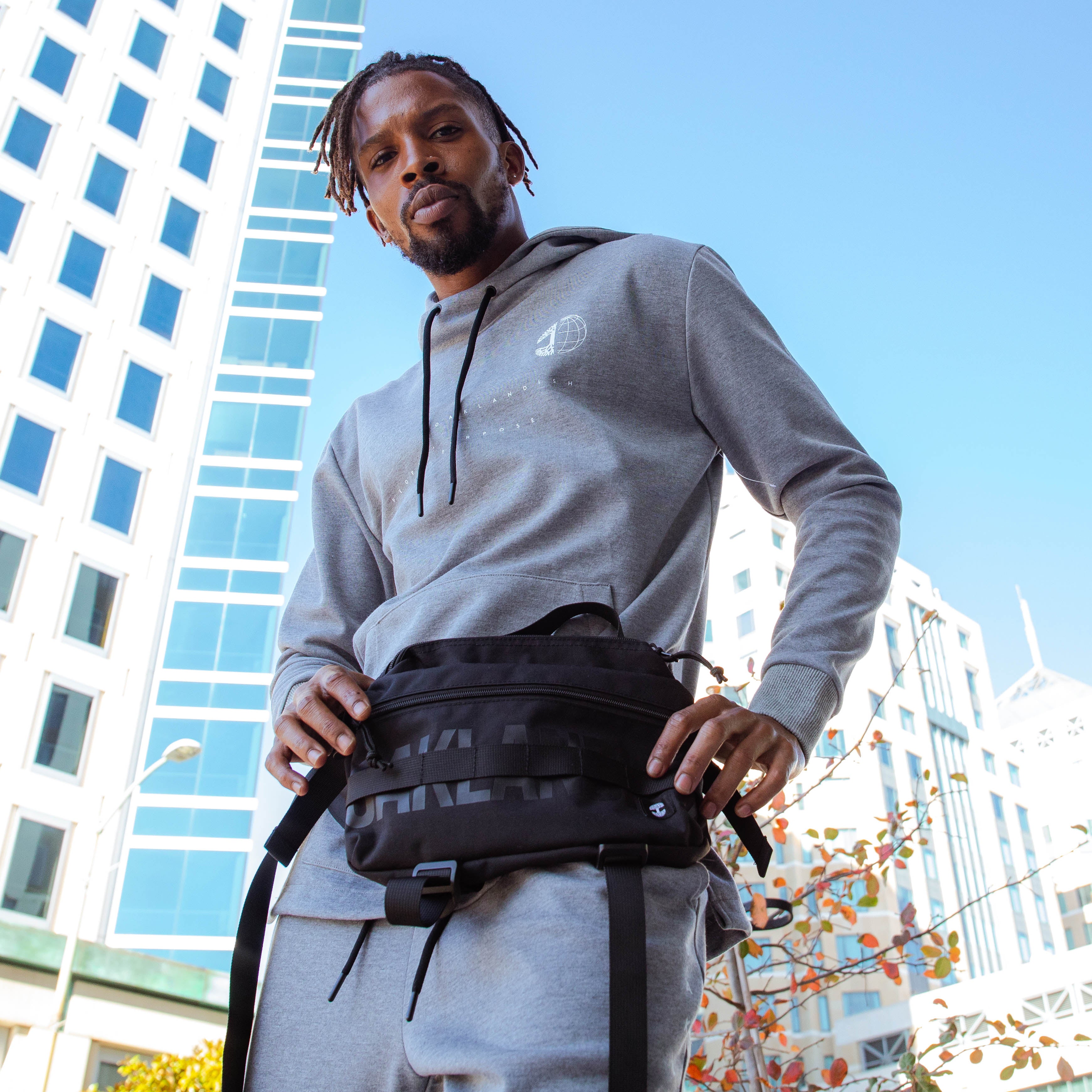 A man standing outside in Oakland holding a black nylon hip bag with a black Oaklandish wordmark & small white Oaklandish tree logo.