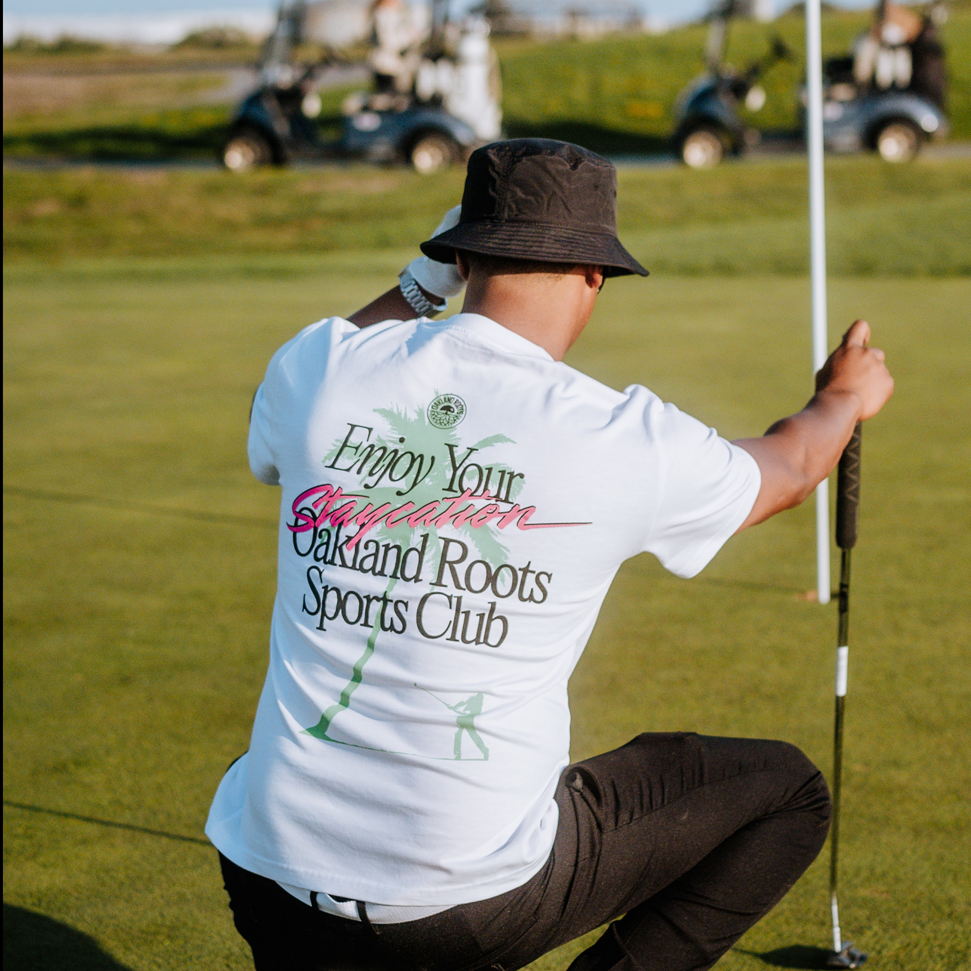 A person is kneeling on a golf course near a flag, holding a golf club. They wear an Oakland Roots SC Staycation Heavy Tee that reads "Enjoy Your Staycation, Oakland Roots Sports Club" and a black bucket hat. In the background, there are golf carts and a distant green landscape promoting social good.