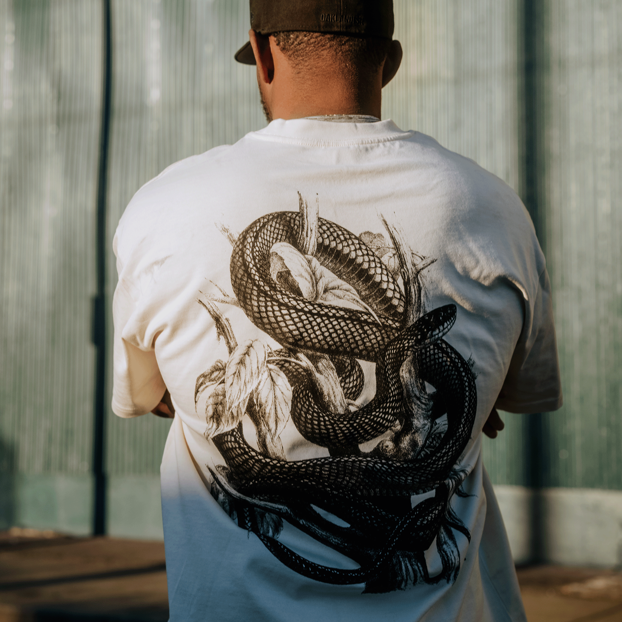 Wearing the Tree Snake Heavy Tee by Oaklandish, a person in men's sizing stands before a corrugated metal wall. The boxy fit, white T-shirt features an intricate black snake print on the back and radiates warmth under the natural sunlight, complemented by a brown baseball cap.