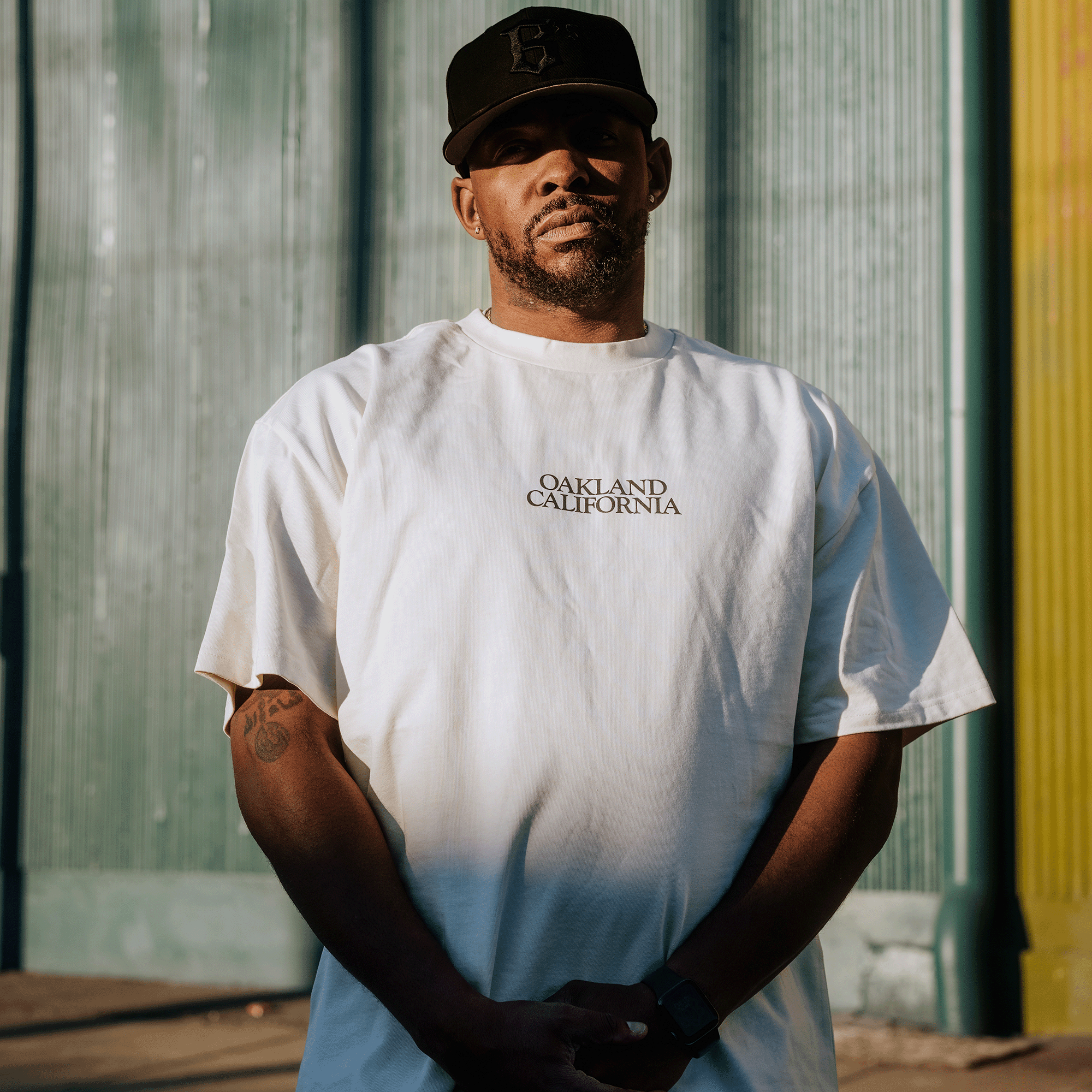 A man stands against a corrugated metal wall with sunlight casting shadows. He wears the "Tree Snake Heavy Tee" from Oaklandish, a heavyweight combed cotton T-shirt with a boxy fit and "Oakland California" text. With a beard and black cap, tattoos on his arms, he clasps his hands, gazing at the camera.