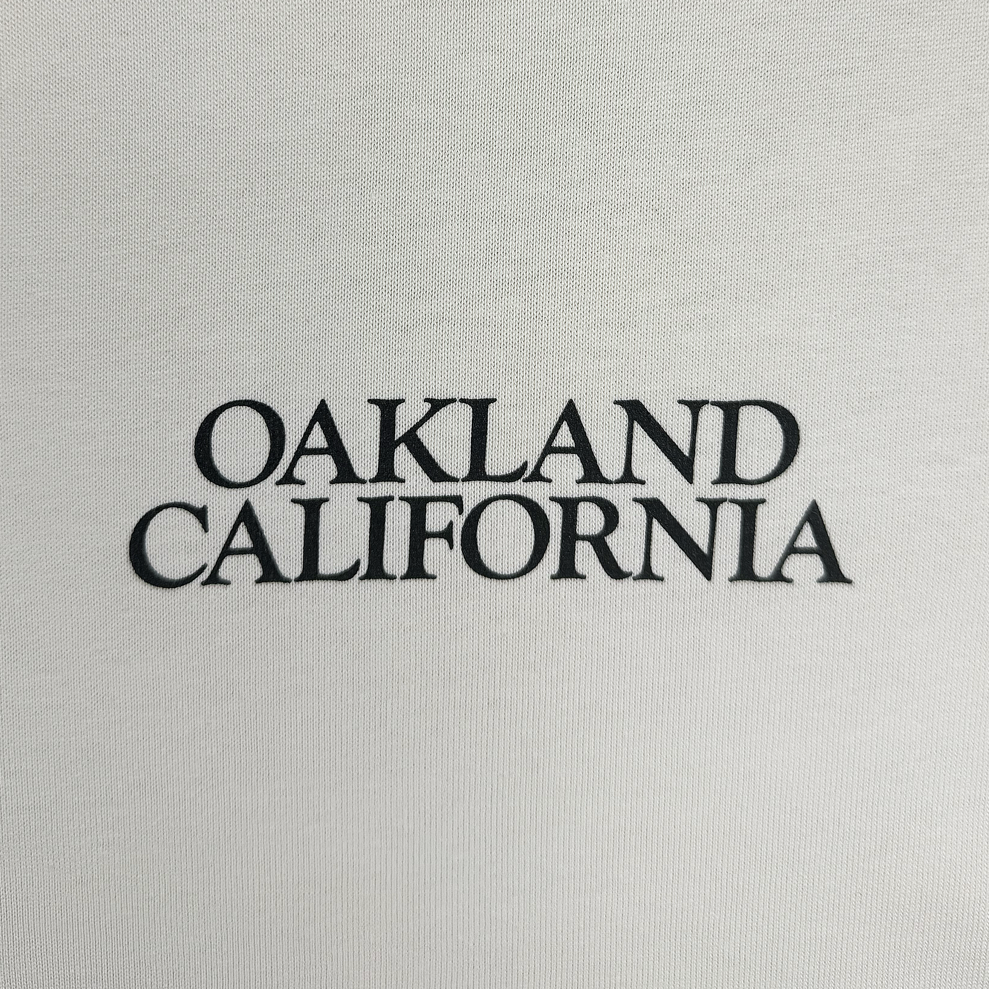 The image features the phrase "OAKLAND CALIFORNIA" in bold, black, uppercase letters centered on a plain white background, echoing the design of a Men's Tree Snake Heavy Tee by Oaklandish. The text's simplicity and clarity shines through on this heavyweight combed cotton boxy fit t-shirt.