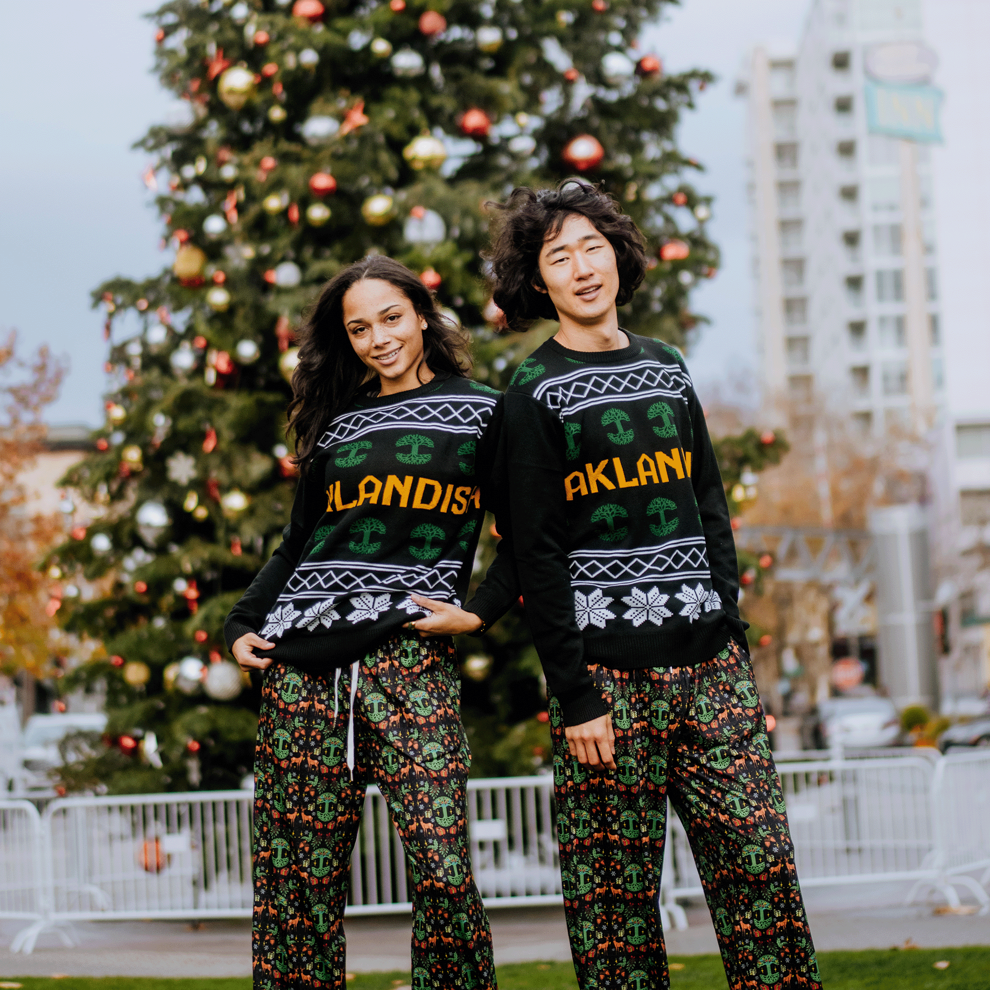 A smiling couple poses outdoors in matching Oaklandish Holiday Pajama Pants and sweaters featuring "Oakland," standing before a large decorated Christmas tree. The unisex fit is clear against a backdrop of city buildings and autumn trees.