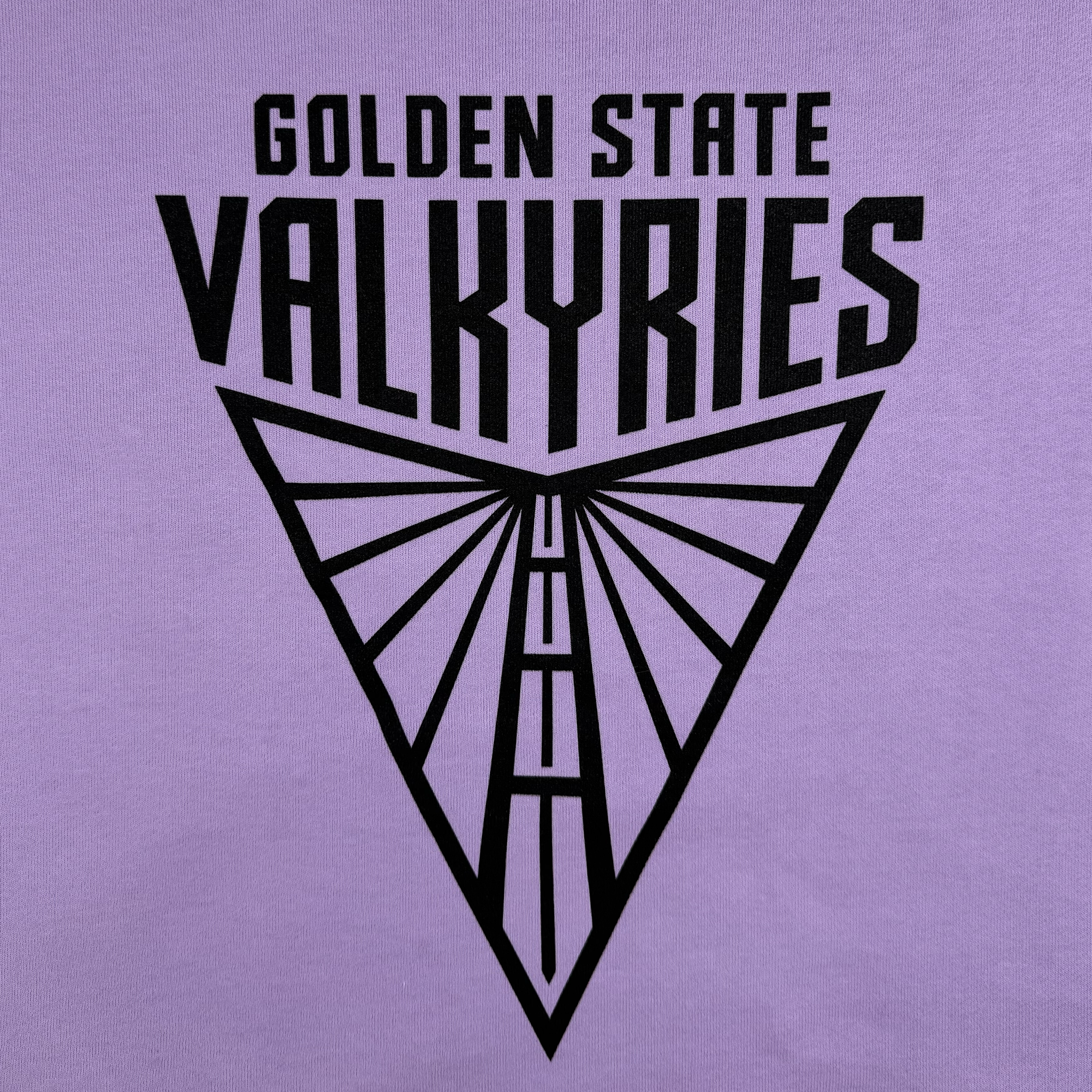 The GSV "Golden State Valkyries Primary Heavy Crewneck" features bold black text above a stylized triangular design with geometric lines resembling a tower, printed on heavyweight preshrunk organic cotton. The background is solid lavender.