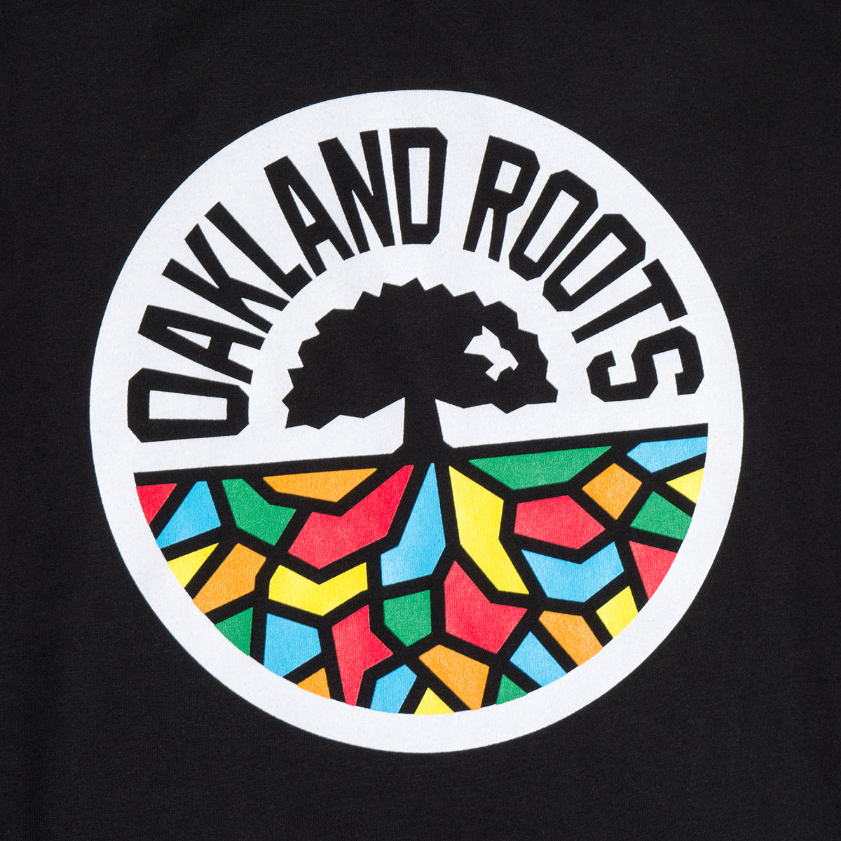 The image features the Women's Oakland Roots SC Classic Tee, a product by Oakland Roots SC. The 100% cotton tee showcases a circular design with "OAKLAND ROOTS" displayed at the top. Below that, there is a black silhouette of a tree, with the ground beneath it depicted in colorful, stained glass-like mosaic pieces. This striking design reflects the team's commitment to social good.