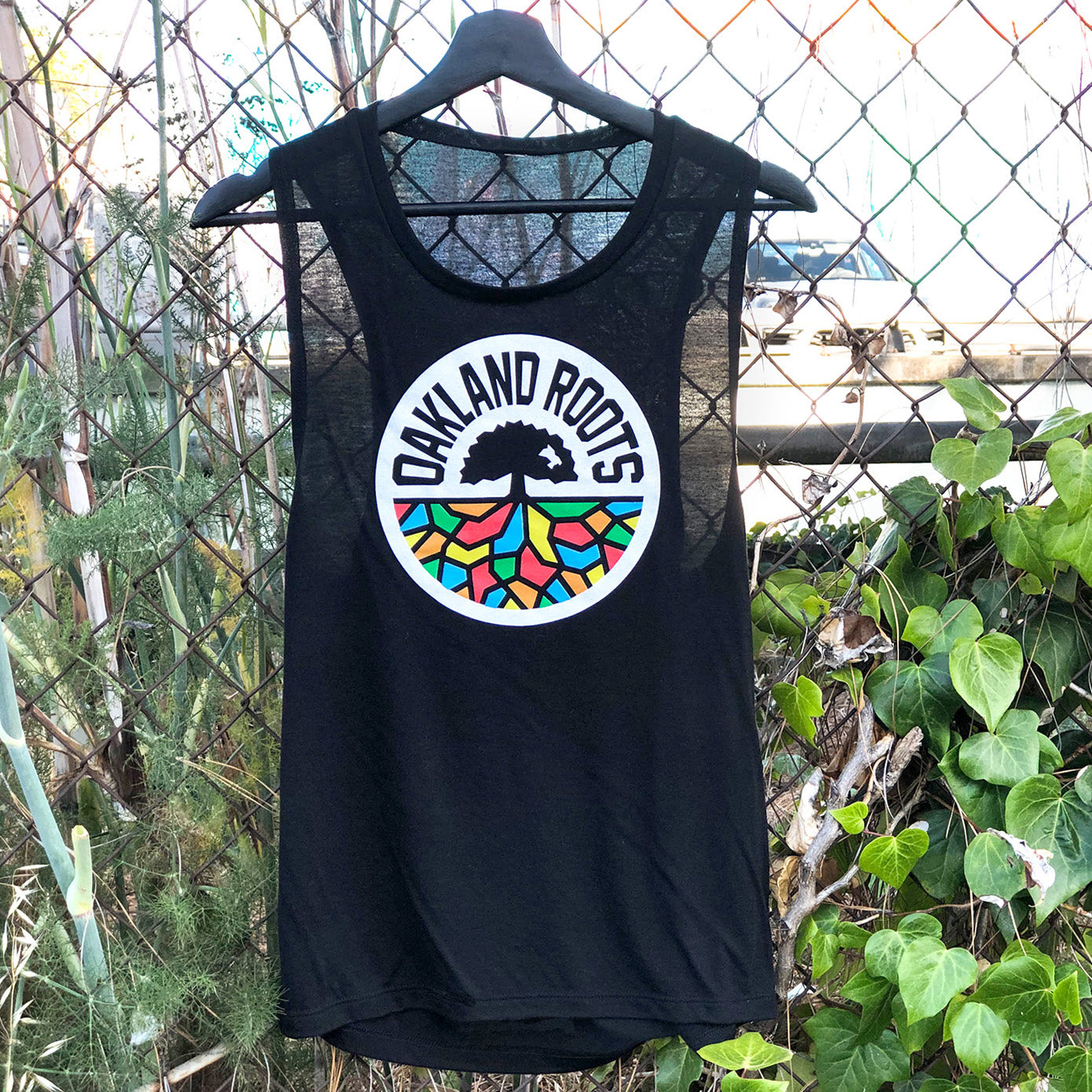 The Women's Oakland Roots SC Logo Tank by Oakland Roots SC hangs on a fence surrounded by greenery. The black tank top prominently displays a colorful circular logo featuring a tree with multicolored roots in red, blue, yellow, and orange, along with the text "OAKLAND ROOTS." A faintly visible structure can be seen behind the fence in the background.