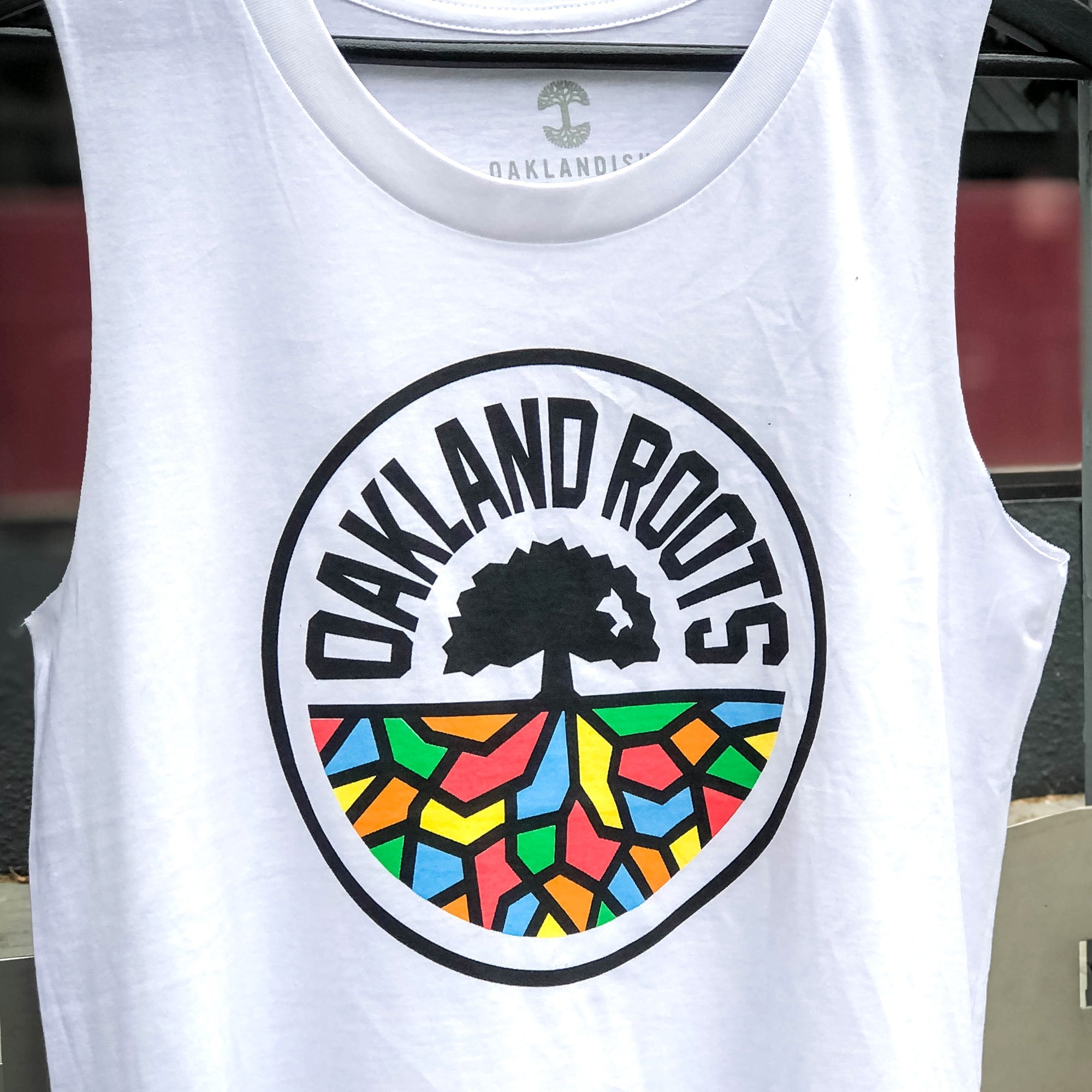 The Women's Oakland Roots SC Logo Tank by Oakland Roots SC is a white tank top that features a circular "OAKLAND ROOTS" logo in the center. The logo depicts a black tree with colorful, geometric soil beneath it in red, blue, yellow, and green. Ideal for fans of the Oakland Roots Sports Club, the background incorporates a blurred view of various colors.