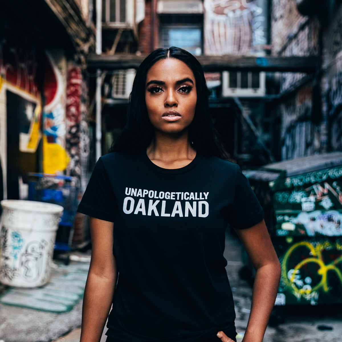 A person with long black hair stands confidently in a graffiti-covered alleyway wearing the 100% cotton Unapologetic Tee by Oaklandish. The vibrant urban backdrop features colorful street art and a barrel adorned with more graffiti.