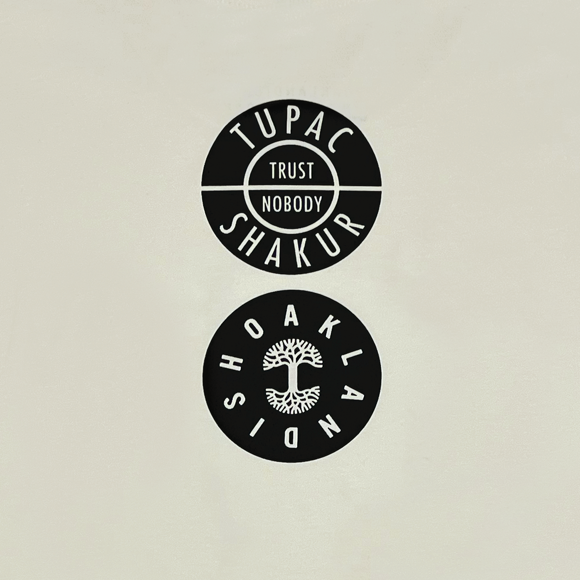 On a light background, there are two round black stickers. The upper sticker embodies the hip-hop spirit with "Wavy Tupac Tee" featuring horizontal lines. Below, the heart of Oaklandish shines with a stylized tree at its center in a symmetrical design.